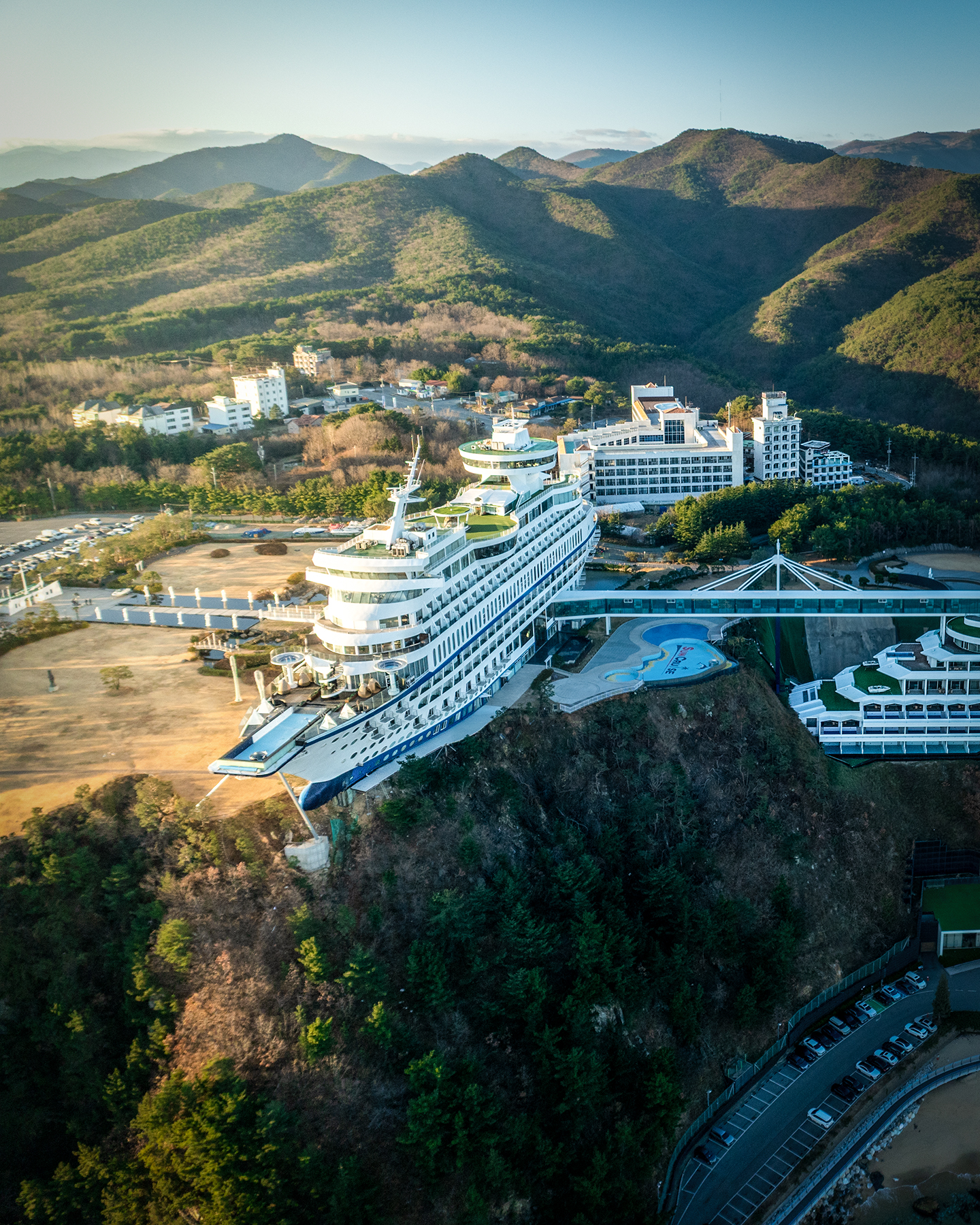 Jeongdongjin - Aerial view shows a large ship-shaped building on a wooded hillside, lit by warm sunlight with layered mountains beyond. - #Jeongdongjin #JeongdongjinBeach #Gangneung #GangneungTravel #GangneungTrip #Korea #SouthKorea #KoreaTravel #KoreaTrip #VisitKorea