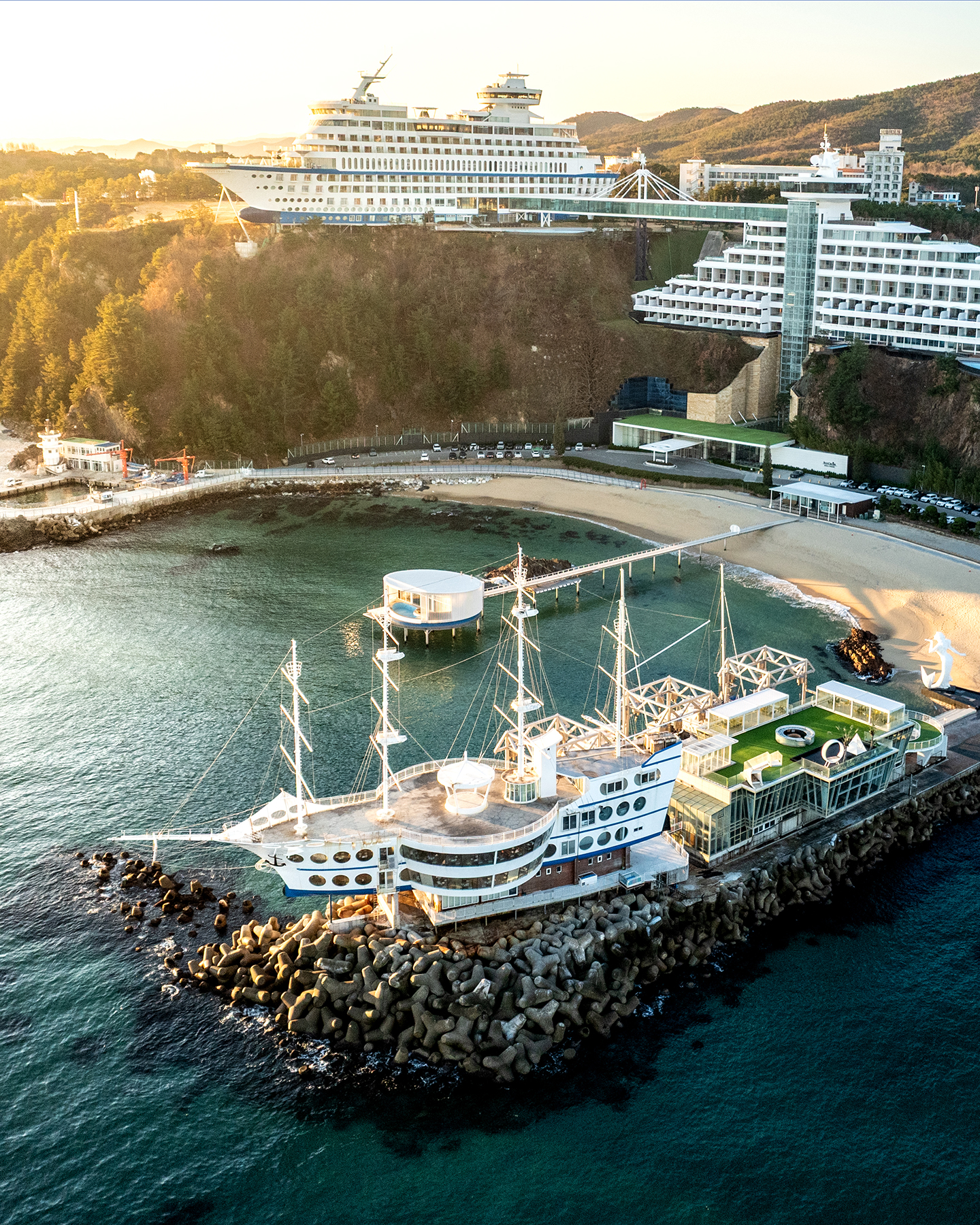 Jeongdongjin - Aerial view of ship-shaped buildings on a rocky coastal breakwater, with a small beach, calm green water, wooded hills, and warm low sunlight. - #Jeongdongjin #JeongdongjinBeach #Gangneung #GangneungTravel #GangneungTrip #Korea #SouthKorea #KoreaTravel #KoreaTrip #VisitKorea
