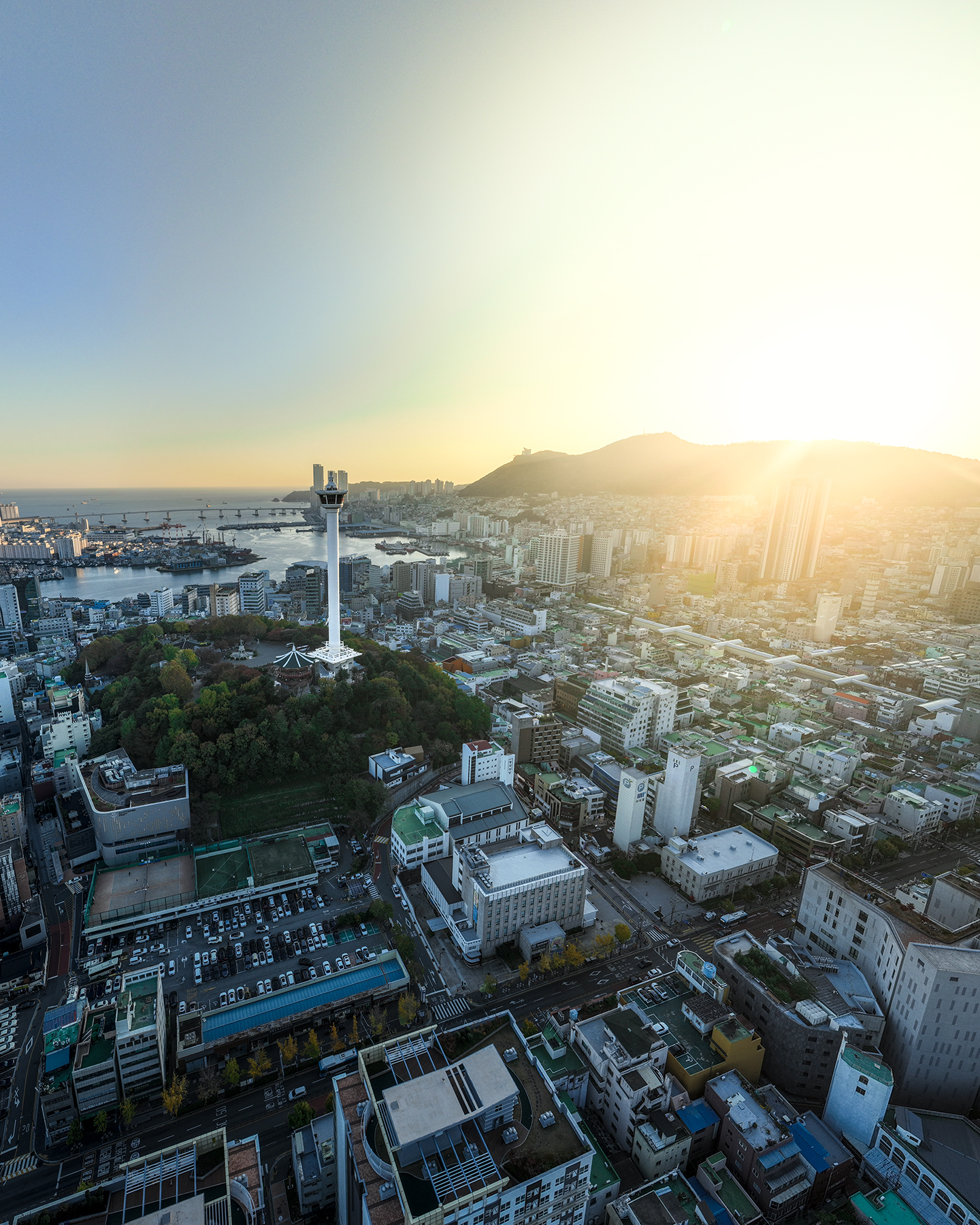 Busan Tower - A white observation tower rises above a dense coastal city as warm sunset light spreads over the harbor and hills. - #BusanTower #YongdusanPark #Busan #BusanKorea #VisitBusan #KoreaTravel #BusanTravel #Cityscape