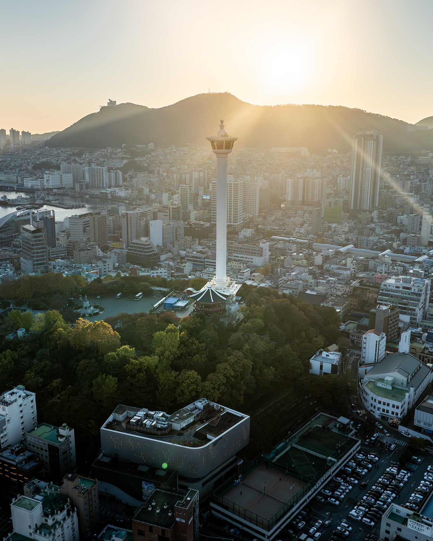 Busan Tower - A tall observation tower rises above a dense cityscape, backlit by warm sunset haze and surrounded by wooded hills. - #BusanTower #YongdusanPark #Busan #BusanKorea #VisitBusan #KoreaTravel #BusanTravel #Cityscape