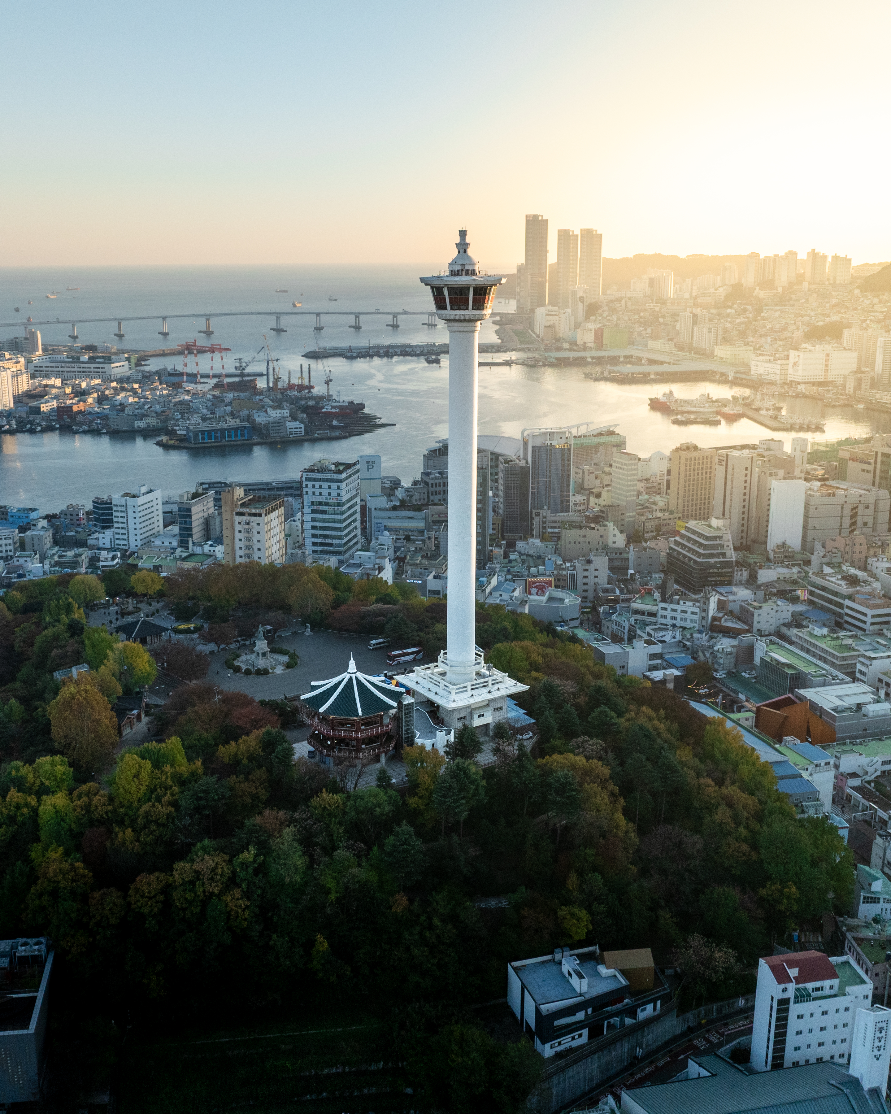 Busan Tower - A white observation tower rises above a coastal city and harbor, bathed in warm hazy sunset light. - #BusanTower #YongdusanPark #Busan #BusanKorea #VisitBusan #KoreaTravel #BusanTravel #Cityscape