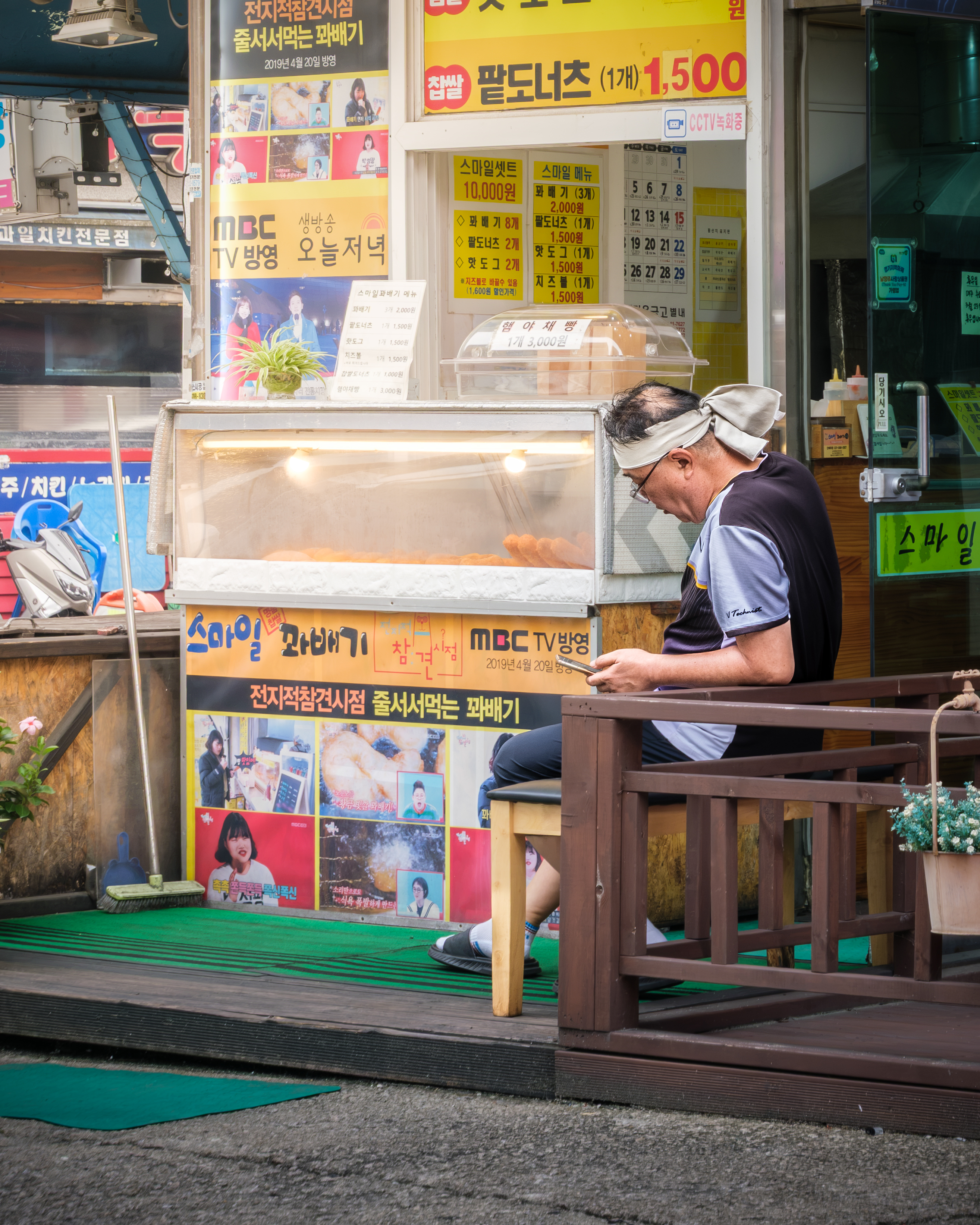 Street - A man looks at his phone beside a brightly signed street snack stall with a glowing food display in daylight. - #Namyangju