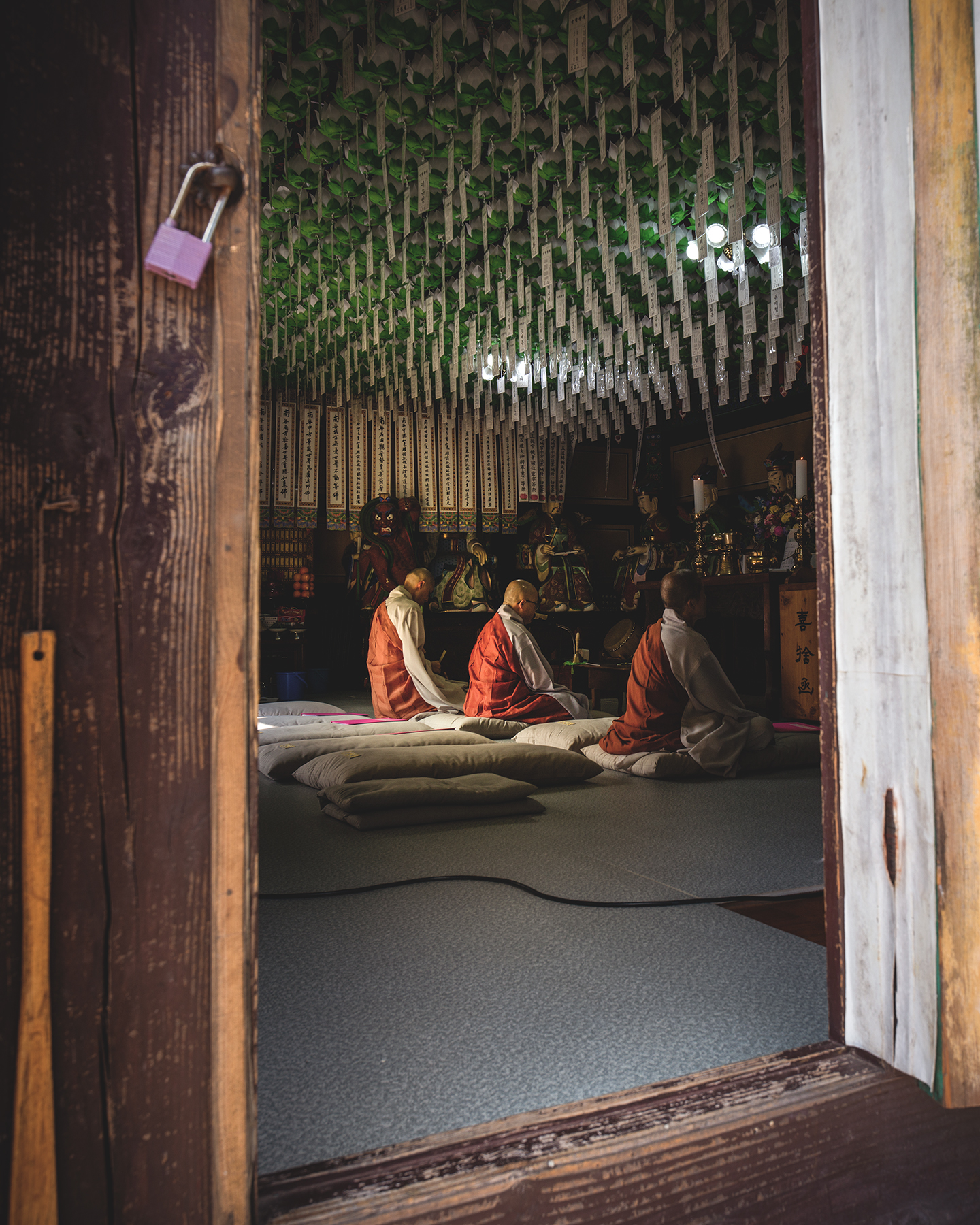 Monk - Three robed monks sit on cushions inside a dim temple hall, framed by a weathered wooden doorway and rows of hanging prayer slips. - #Buddhism #Buddhist #BuddhistMonk #BuddhistMonks #BuddhistTemple #BuddhistCulture #BuddhistTradition #KoreanBuddhism #KoreanTemple #TempleLife