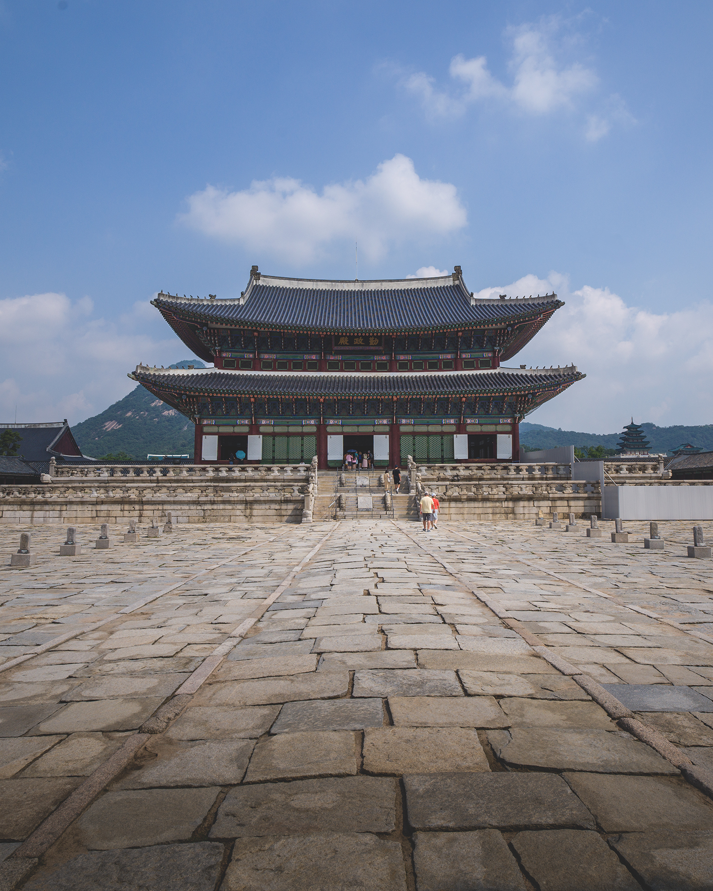 Gyeongbokgung - A traditional palace hall stands symmetrically beyond a wide stone courtyard, with mountains and soft clouds under bright daylight. - #Gyeongbokgung #GyeongbokgungPalace #Seoul #SeoulKorea #SouthKorea #KoreaTravel #VisitKorea #VisitSeoul #KoreaPhotography #SeoulPhotography
