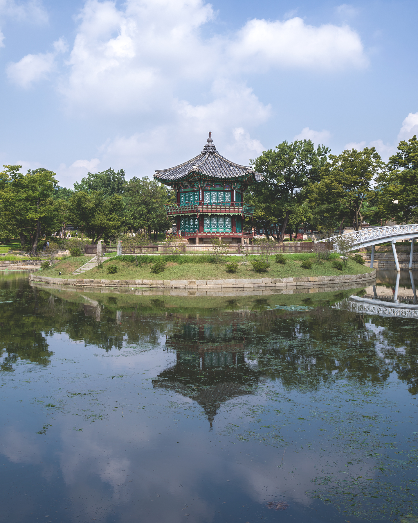 Gyeongbokgung - A traditional pavilion stands on a tree-lined island, reflected in a calm pond under a bright sky with soft clouds. - #Gyeongbokgung #GyeongbokgungPalace #Seoul #SeoulKorea #SouthKorea #KoreaTravel #VisitKorea #VisitSeoul #KoreaPhotography #SeoulPhotography
