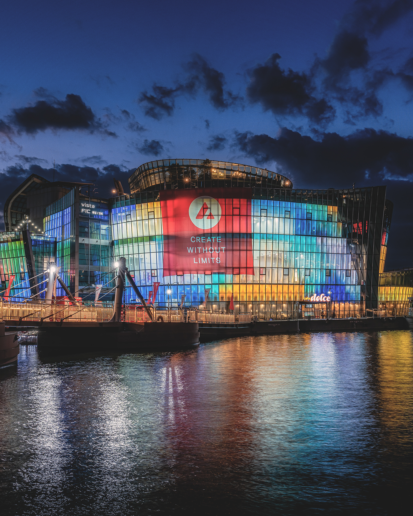 Sebitseom - A colorful glass riverside building glows at night beneath dark clouds, with bright reflections rippling across the water. - #Sebitseom #SebitIsland #SebitFloatingIsland #HanRiver #Hangang #Seoul #SeoulKorea #SouthKorea #KoreaTravel #SeoulTravel