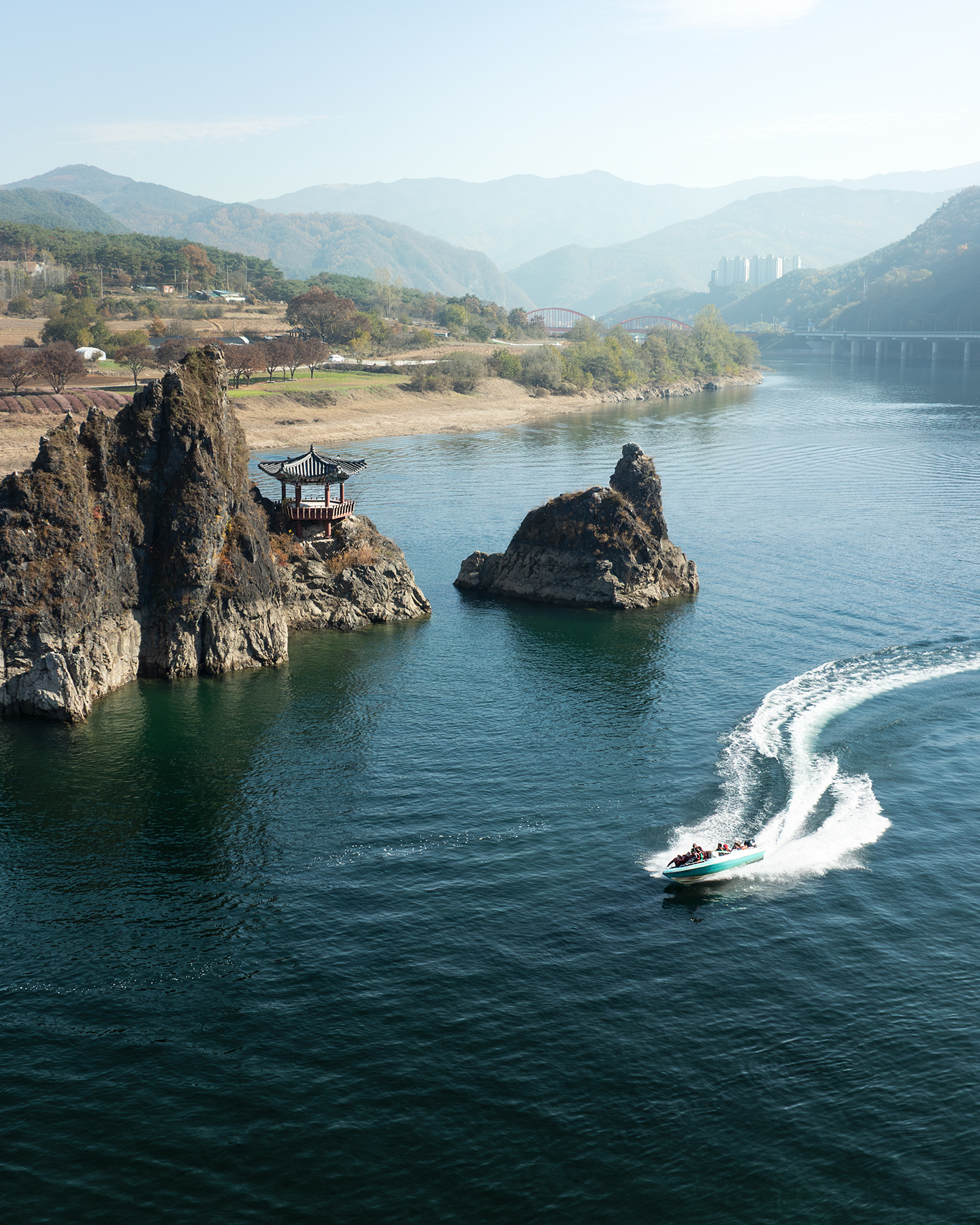 Dodamsambong - A speedboat carves a bright wake past rocky river islets with a small pavilion, backed by hazy mountains and bridges. - #Dodamsambong #DodamSambongPeaks #Danyang #Chungcheongbukdo #SouthKorea #KoreaTravel #VisitKorea #DanyangTravel #KoreaPhotography #TravelPhotography