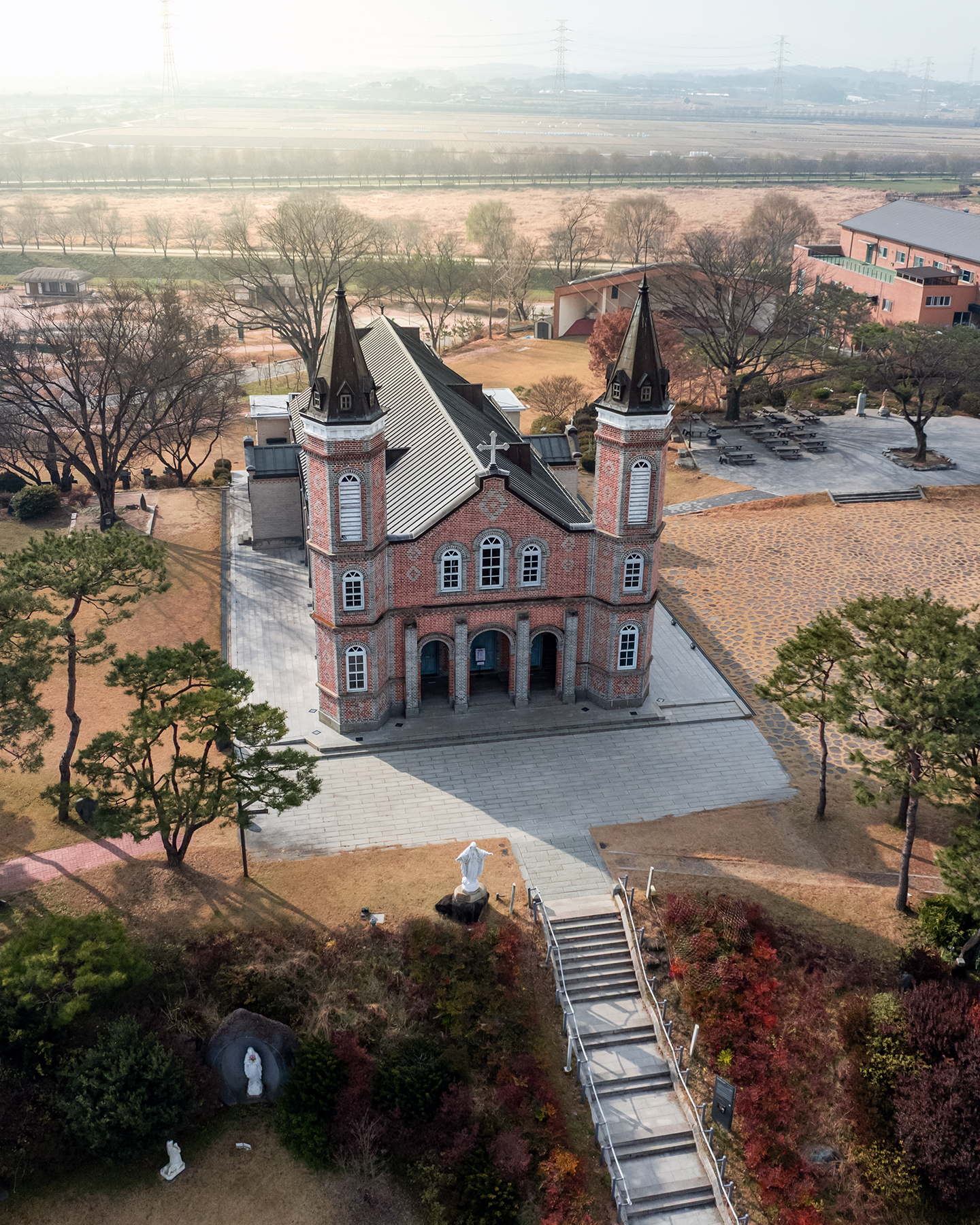 Hapdeok Catholic Church - An aerial view shows a red-brick church with twin spires, a front statue, bare trees, and hazy fields beyond. - #HapdeokCatholicChurch #HapdeokChurch #HapdeokCathedral #Dangjin #Chungcheongnamdo #SouthKorea #KoreaTravel #KoreaPhotography #TravelPhotography #ChurchPhotograph