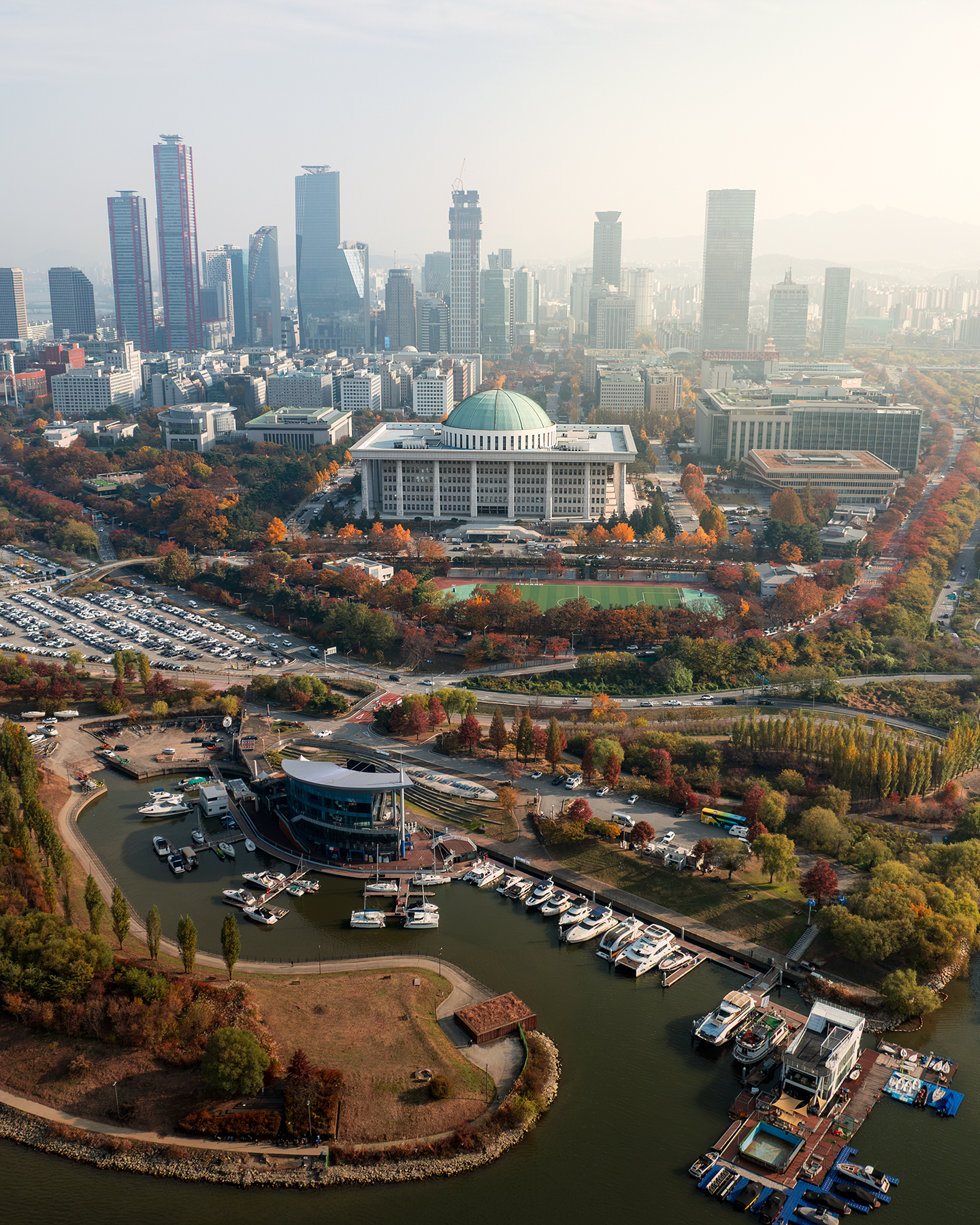 Yeouido - An aerial cityscape shows a domed building, marina, high-rises, and autumn trees under soft hazy light. - #Yeouido #NationalAssembly #NationalAssemblyOfKorea #KoreanNationalAssembly #NationalAssemblyBuilding #Seoul #SeoulKorea #SouthKorea #KoreaTravel #SeoulTravel