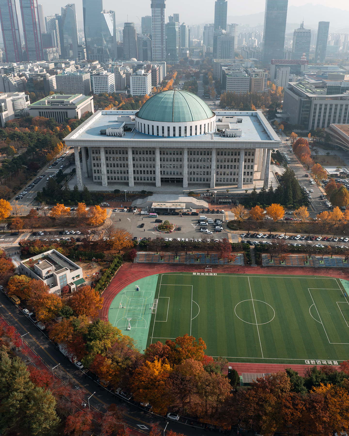 Yeouido - An aerial view shows a large domed building, autumn trees, city towers, roads, and a sports field under hazy daylight. - #Yeouido #NationalAssembly #NationalAssemblyOfKorea #KoreanNationalAssembly #NationalAssemblyBuilding #Seoul #SeoulKorea #SouthKorea #KoreaTravel #SeoulTravel