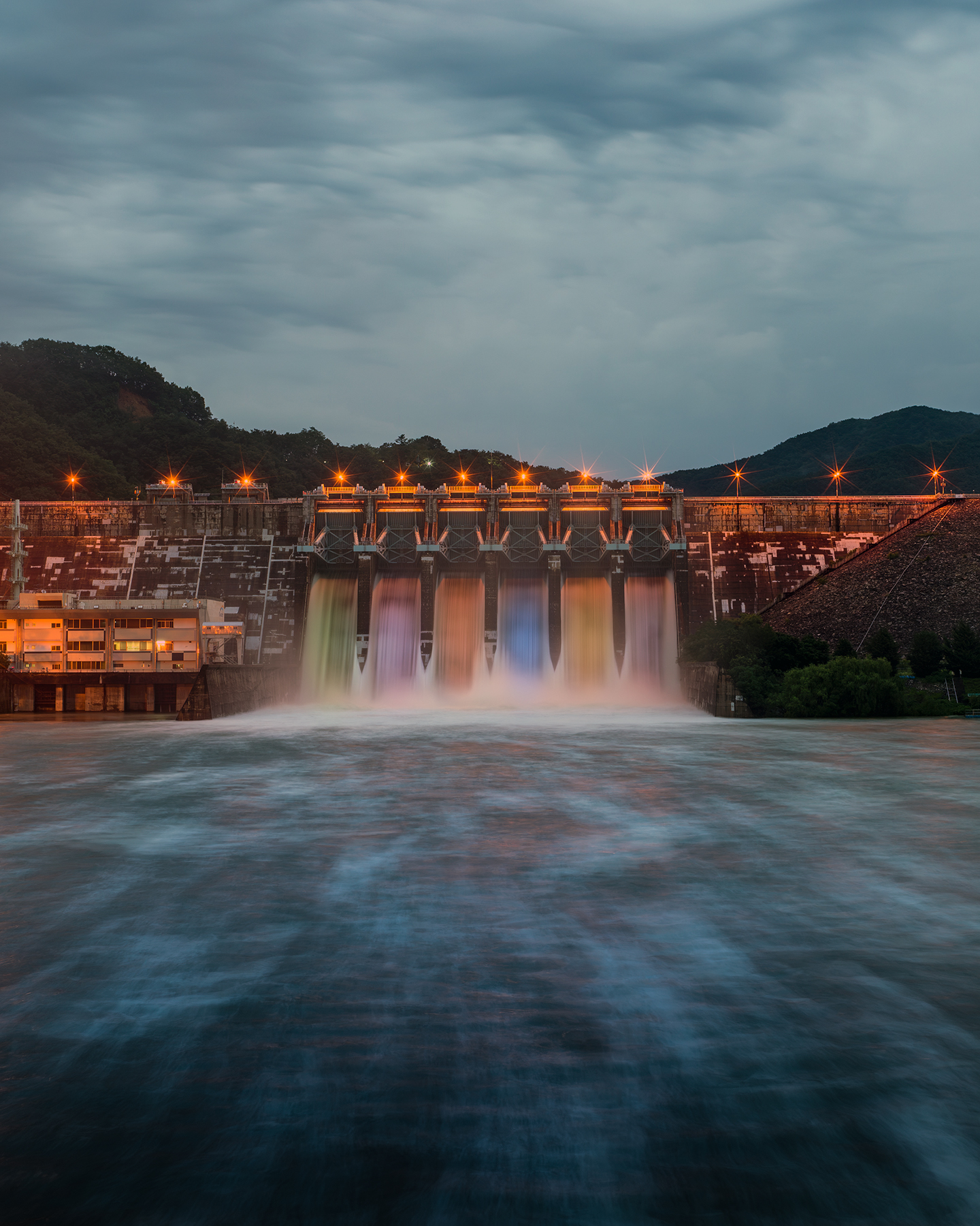 Daecheong Dam - A lit dam releases colorful streams into a wide river beneath a cloudy evening sky, framed by dark hills. - #DaecheongDam #DaecheongLake #Dam #Spillway #WaterRelease #DamRelease #LongExposure #LongExposurePhotography #LandscapePhotography #NightPhotography