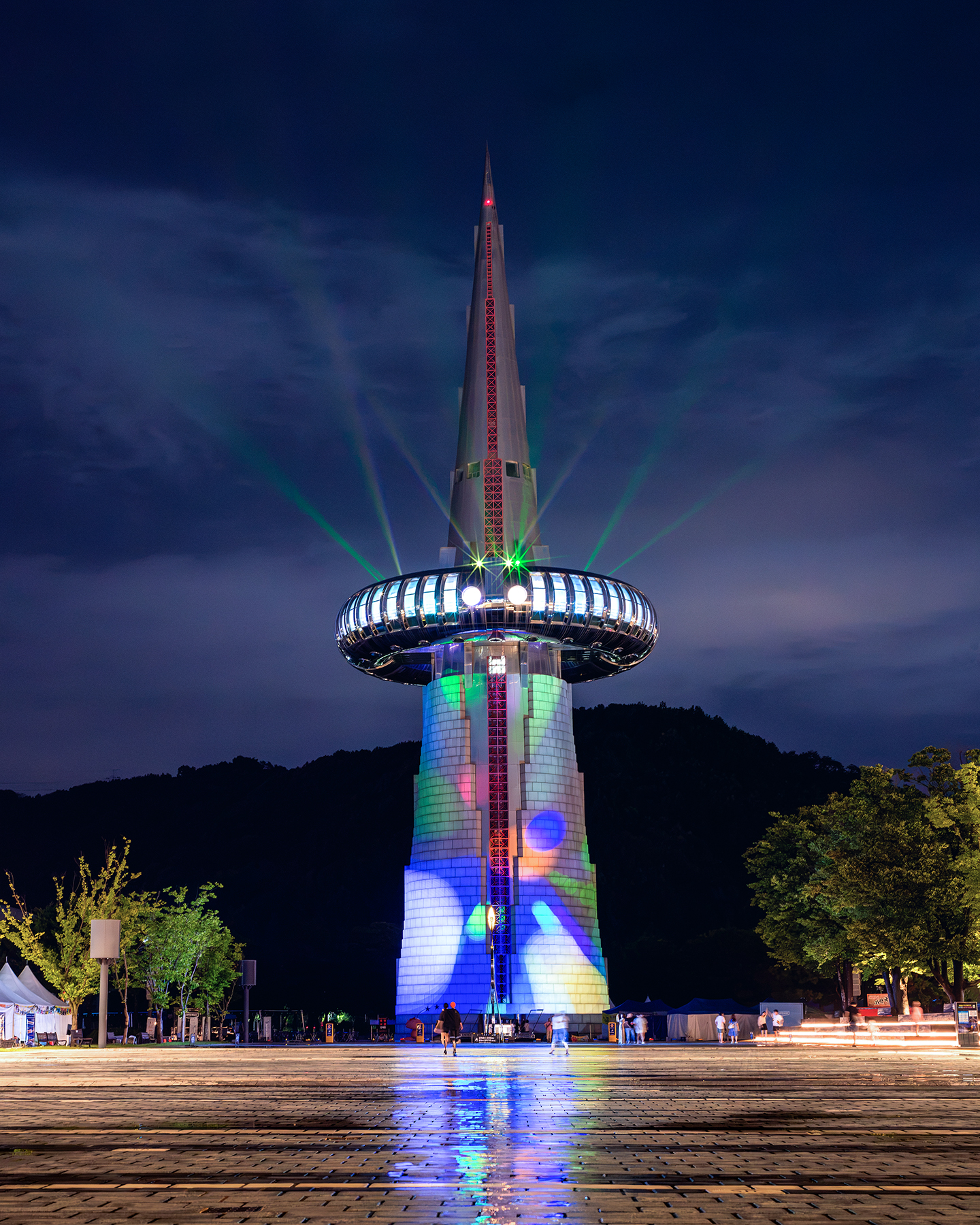 Hanbit Tower - A tall illuminated tower rises over a wet plaza at night, with colorful projections and green beams against a dark sky. - #HanbitTower #Daejeon #DaejeonExpo #ExpoSciencePark #ExpoPark #DaejeonLandmark #Nightscape #NightPhotography #Cityscape #CityLights