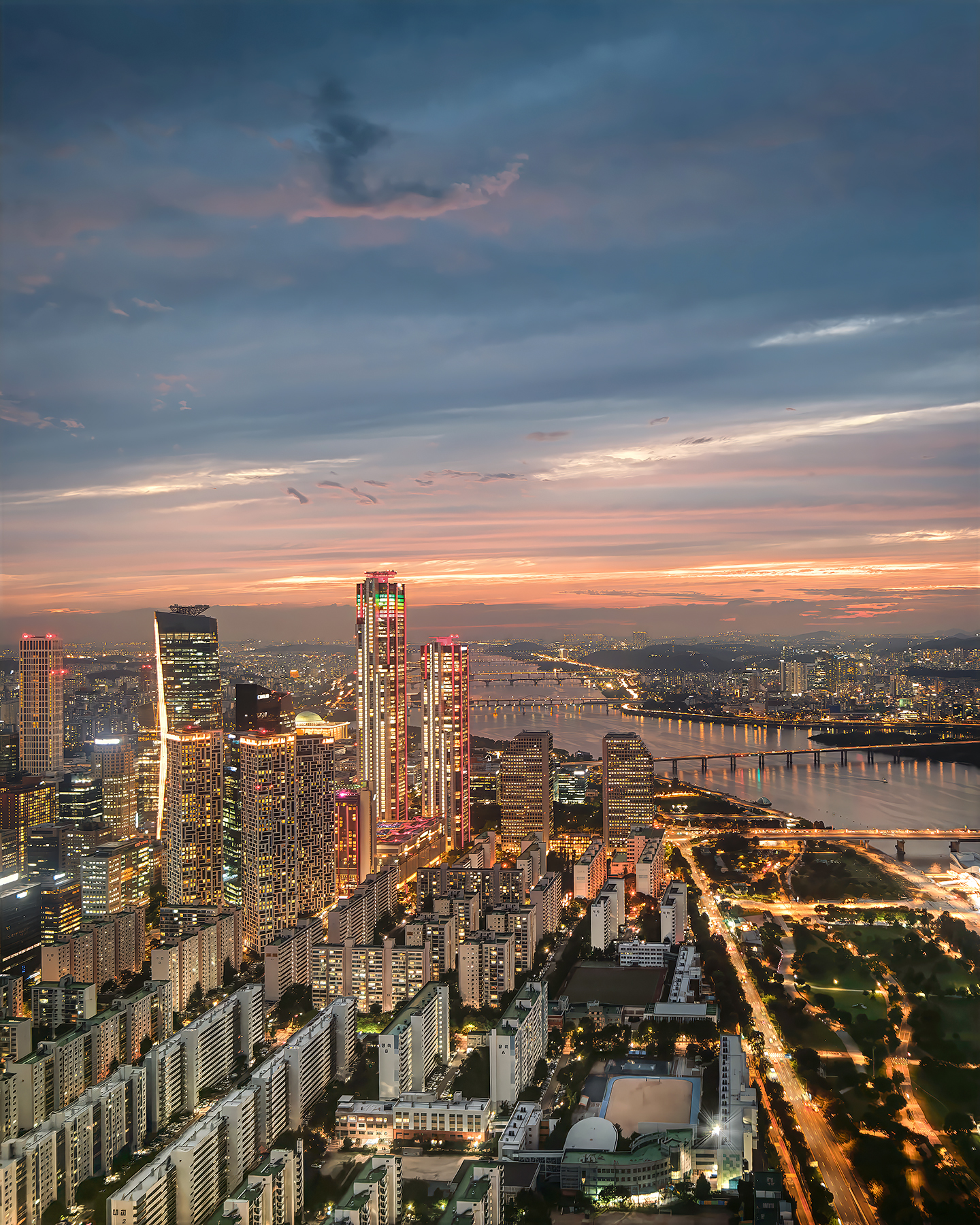 Yeouido - A glowing city skyline rises beside a broad river at dusk, with bridges, traffic lights, and layered clouds under a warm sunset. - #Seoul #SeoulKorea #SouthKorea #Yeouido #YeouidoSkyline #SeoulSkyline #SeoulCityscape #HanRiver #HanRiverView #SeoulNightView