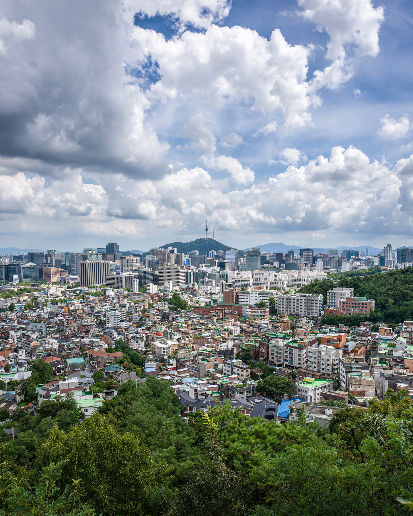 Namsan - A dense city skyline stretches toward a green hill with a tower under towering cumulus clouds and bright midday light. - #Inwangsan #Namsan #NamsanSeoulTower #SeoulTower #SeoulSkyline #SeoulView #SeoulCityscape #SeoulLandscape #SeoulMountains #SeoulPhotography