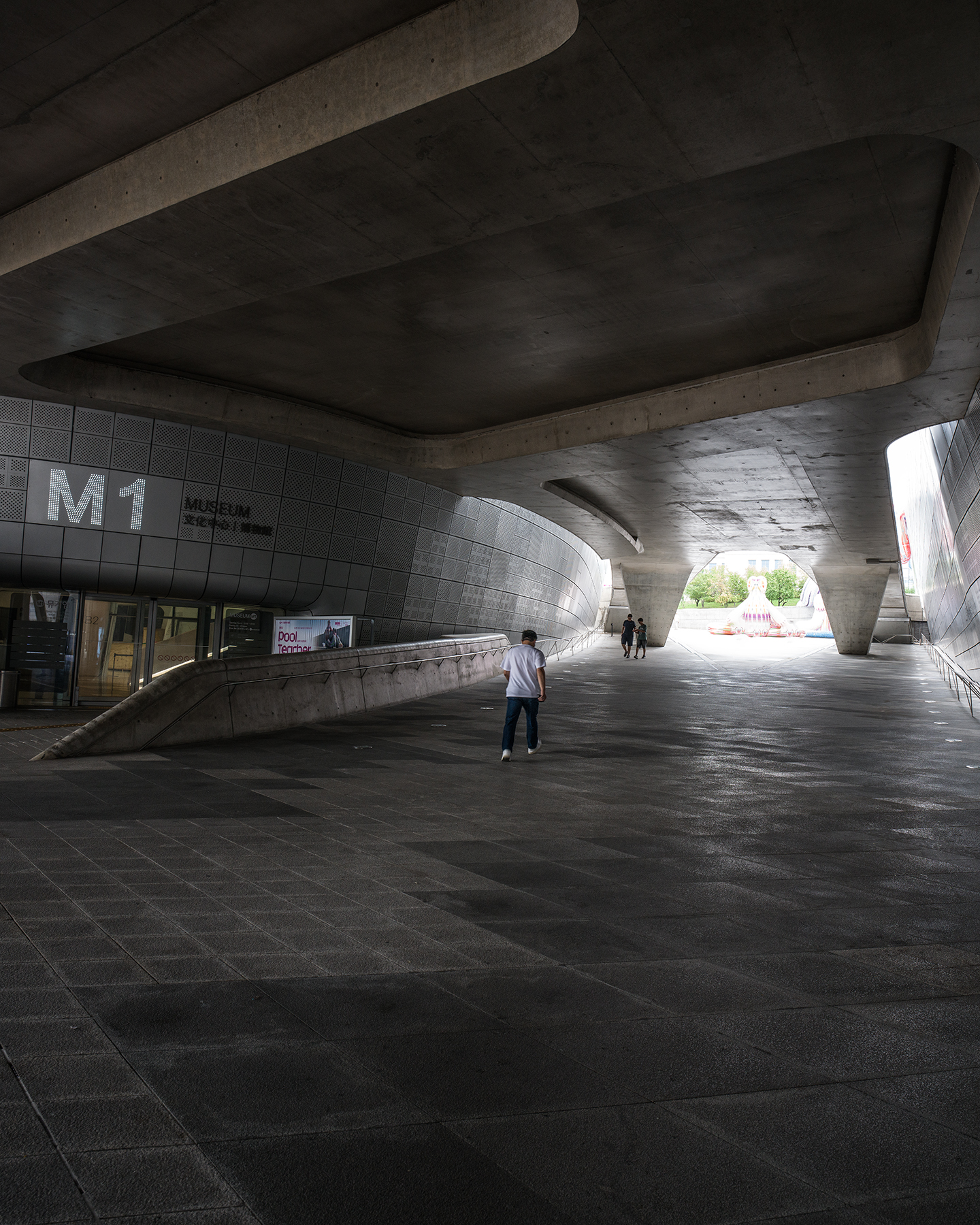 DDP - Pedestrians walk through a shadowed concrete passage beneath sweeping curved architecture toward bright daylight. - #DDP #DongdaemunDesignPlaza #Dongdaemun #Seoul #SeoulKorea #VisitSeoul #SeoulTravel #KoreaTravel #SeoulArchitecture #ModernArchitecture