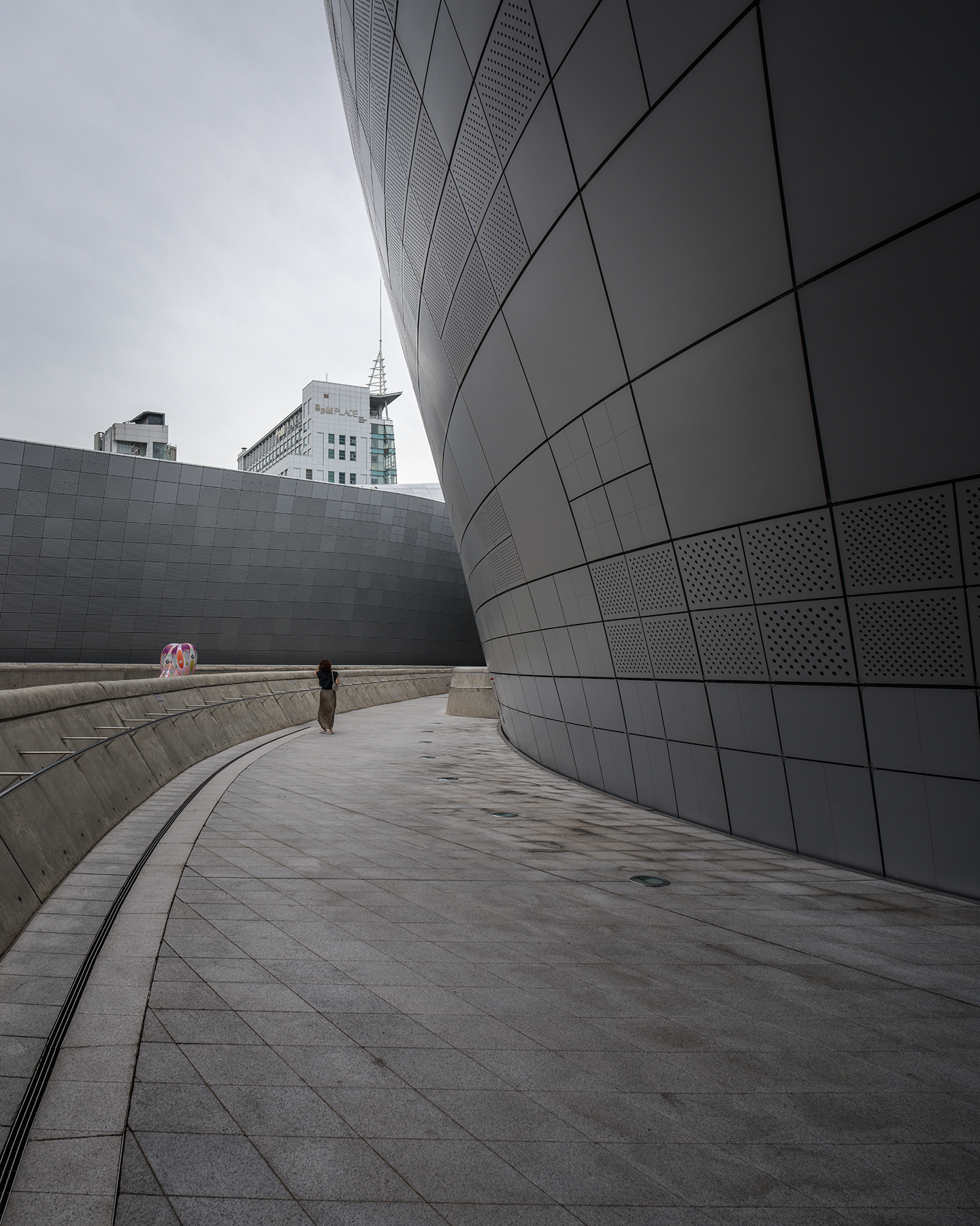 DDP - A lone pedestrian walks along a sweeping curved plaza beside dark metal architectural panels under an overcast sky. - #DDP #DongdaemunDesignPlaza #Dongdaemun #Seoul #SeoulKorea #VisitSeoul #SeoulTravel #KoreaTravel #SeoulArchitecture #ModernArchitecture