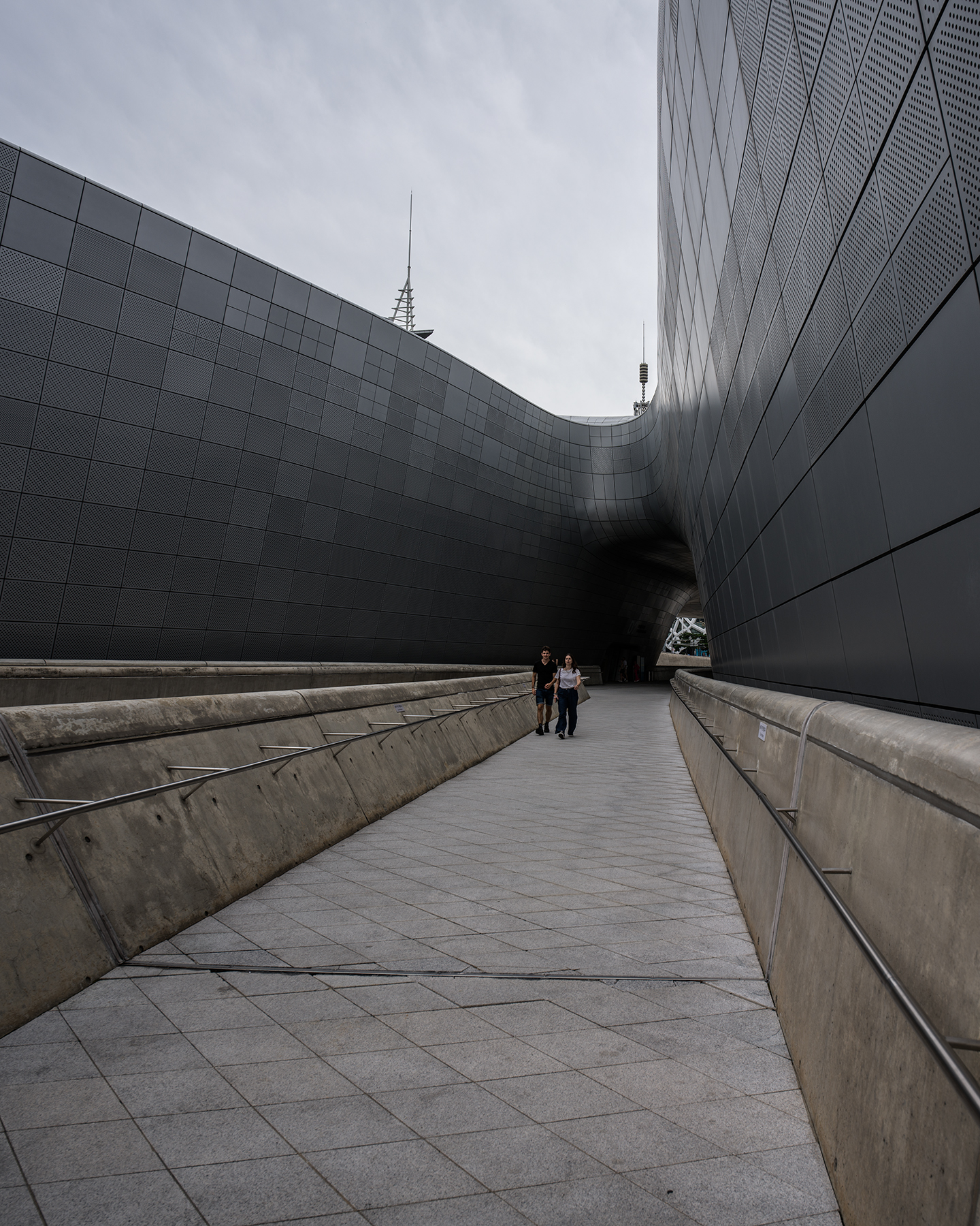 DDP - Two pedestrians walk along a wide tiled ramp between sweeping gray metal walls under an overcast sky. - #DDP #DongdaemunDesignPlaza #Dongdaemun #Seoul #SeoulKorea #VisitSeoul #SeoulTravel #KoreaTravel #SeoulArchitecture #ModernArchitecture