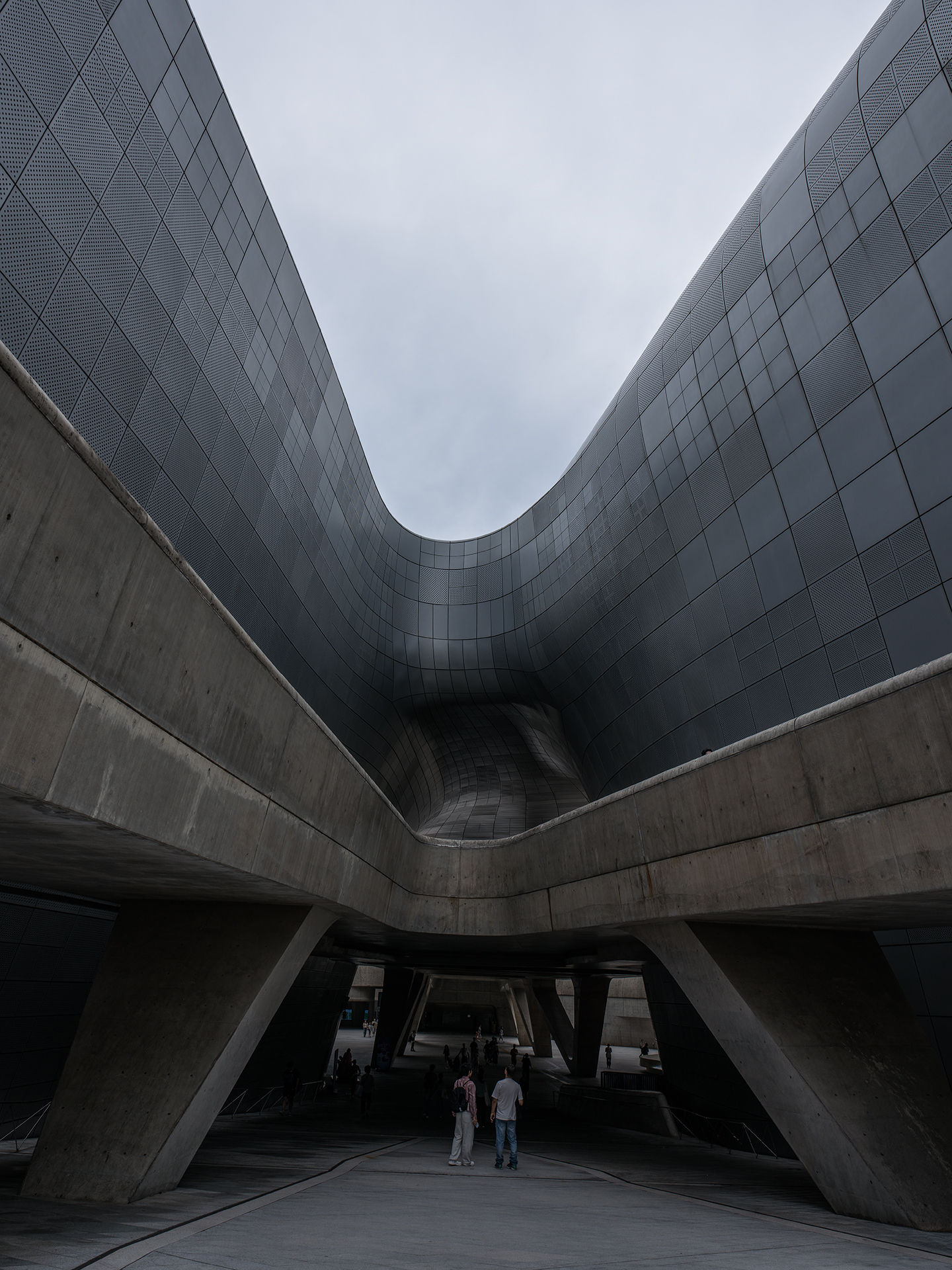 DDP - Curving metal facades and concrete supports frame a shadowed pedestrian space beneath an overcast sky. - #DDP #DongdaemunDesignPlaza #Dongdaemun #Seoul #SeoulKorea #VisitSeoul #SeoulTravel #KoreaTravel #SeoulArchitecture #ModernArchitecture