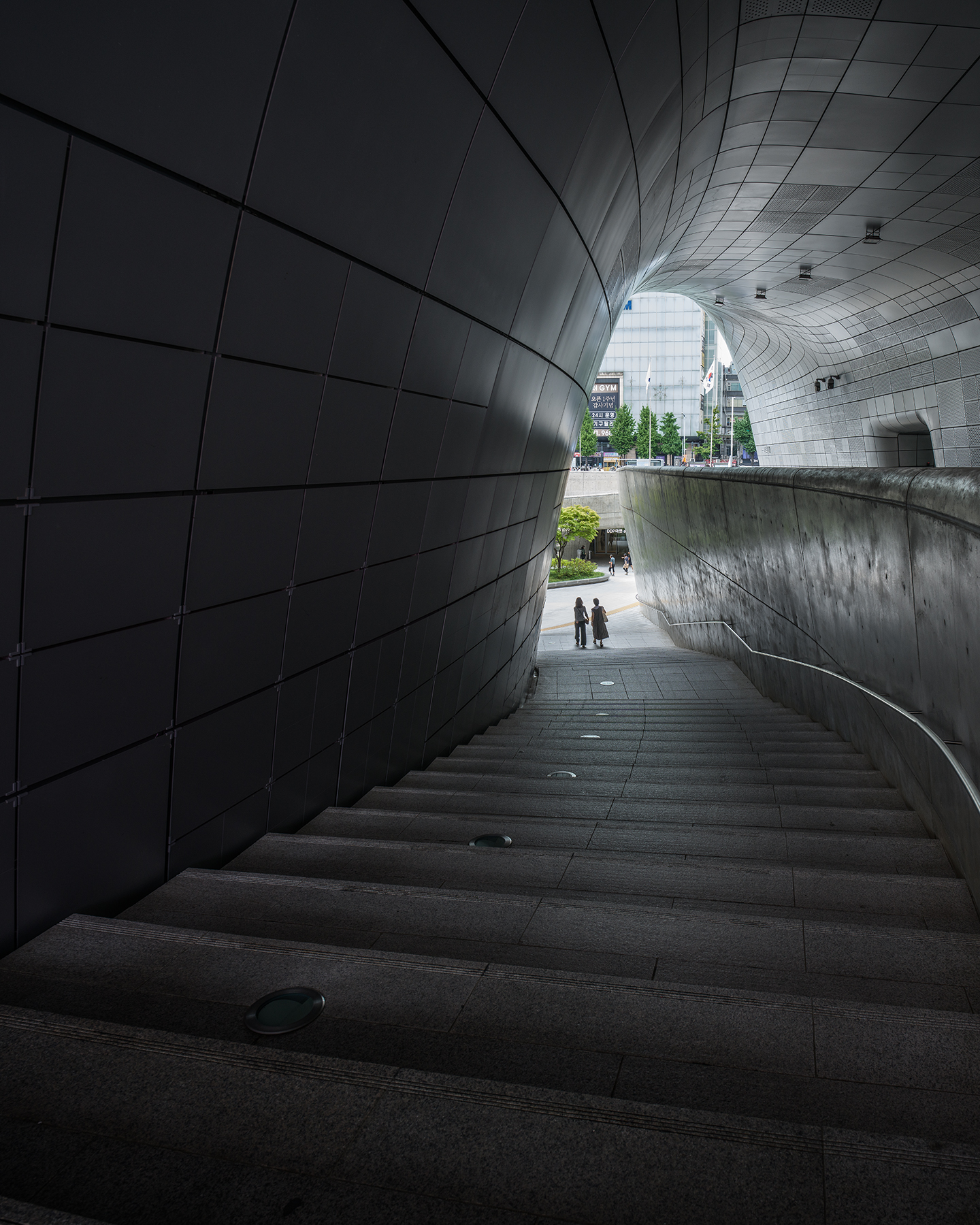DDP - A dim stairway descends between sweeping metal-clad walls toward a bright urban opening with small pedestrians. - #DDP #DongdaemunDesignPlaza #Dongdaemun #Seoul #SeoulKorea #VisitSeoul #SeoulTravel #KoreaTravel #SeoulArchitecture #ModernArchitecture