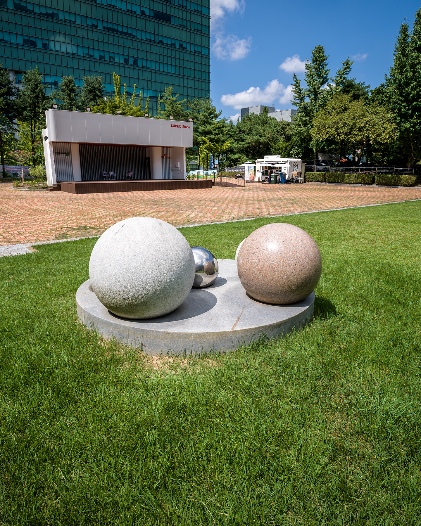 Park - Spherical sculptures sit on a grassy park lawn beside a small stage, trees, and a glass building under bright midday sun. - #Cheongju #CheongjuPark #CheongjuCity #KoreaPark #KoreanPark #ParkInKorea #UrbanPark #CityPark #NatureInKorea #KoreaTravel