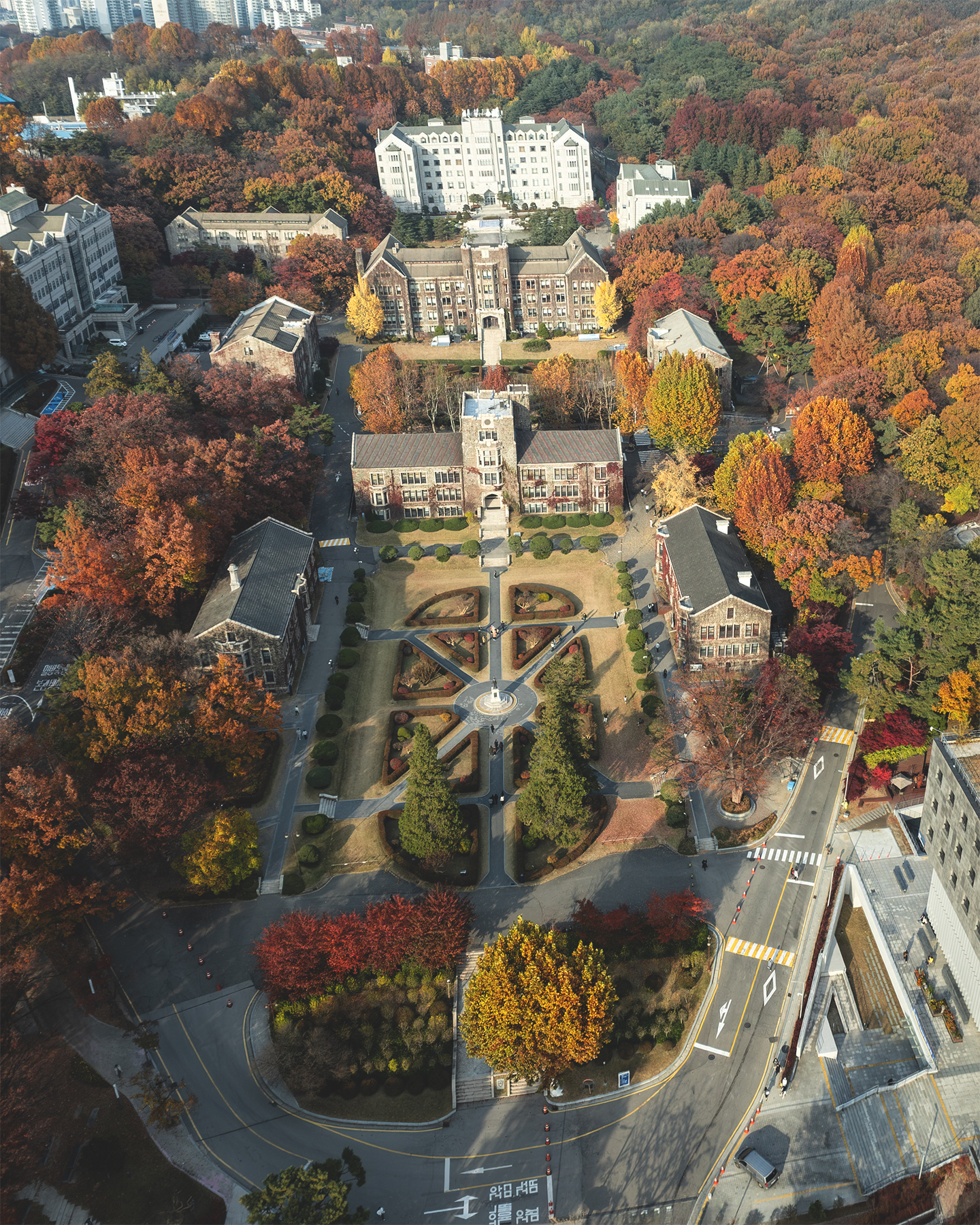 Yonsei - An aerial view shows historic stone campus buildings, formal gardens, roads, and dense autumn trees in warm daylight. - #YonseiUniversity #Yonsei #YonseiUniversityCampus #YonseiInAutumn #AutumnAtYonsei #AutumnCampus #AutumnInSeoul #FallColors #AutumnColors #FallFoliage