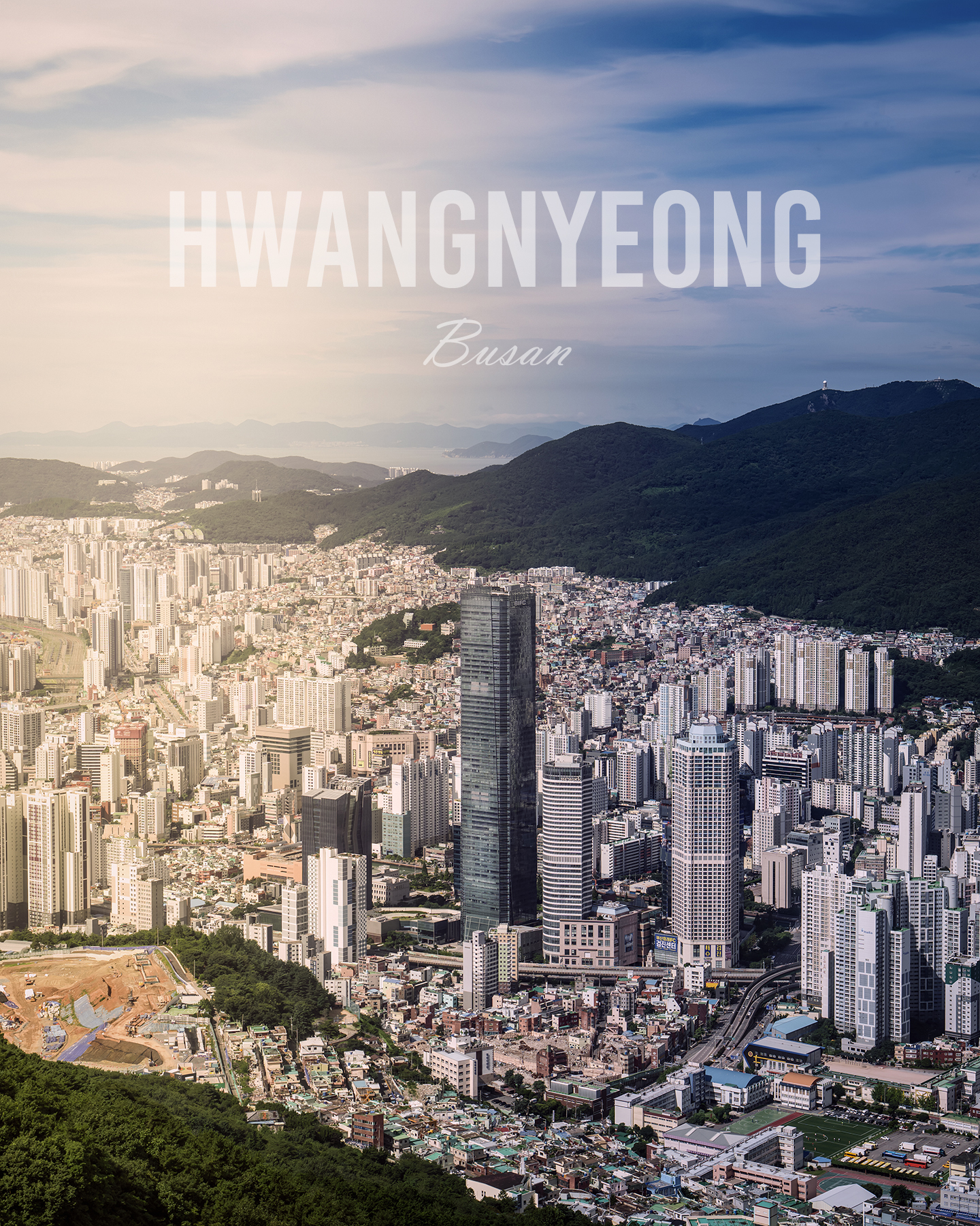 Hwangnyeongsan - A dense city skyline spreads between green mountains under soft evening light and a hazy blue sky. - #Hwangnyeongsan #HwangnyeongMountain #MunhyeonDong #BIFC #BusanInternationalFinanceCenter #Busan #BusanSkyline #BusanCityscape #BusanNightView #BusanNightscape