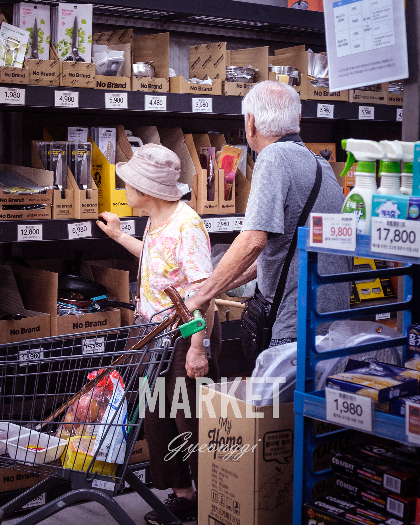 Market - Two older shoppers browse kitchenware on crowded supermarket shelves beside a cart under bright indoor retail lighting. - #Shopping #GroceryShopping #Supermarket #NoBrand #EverydayLife #DailyLife #KoreanLifestyle #SeniorLife #ElderlyCouple #ShoppingTogether