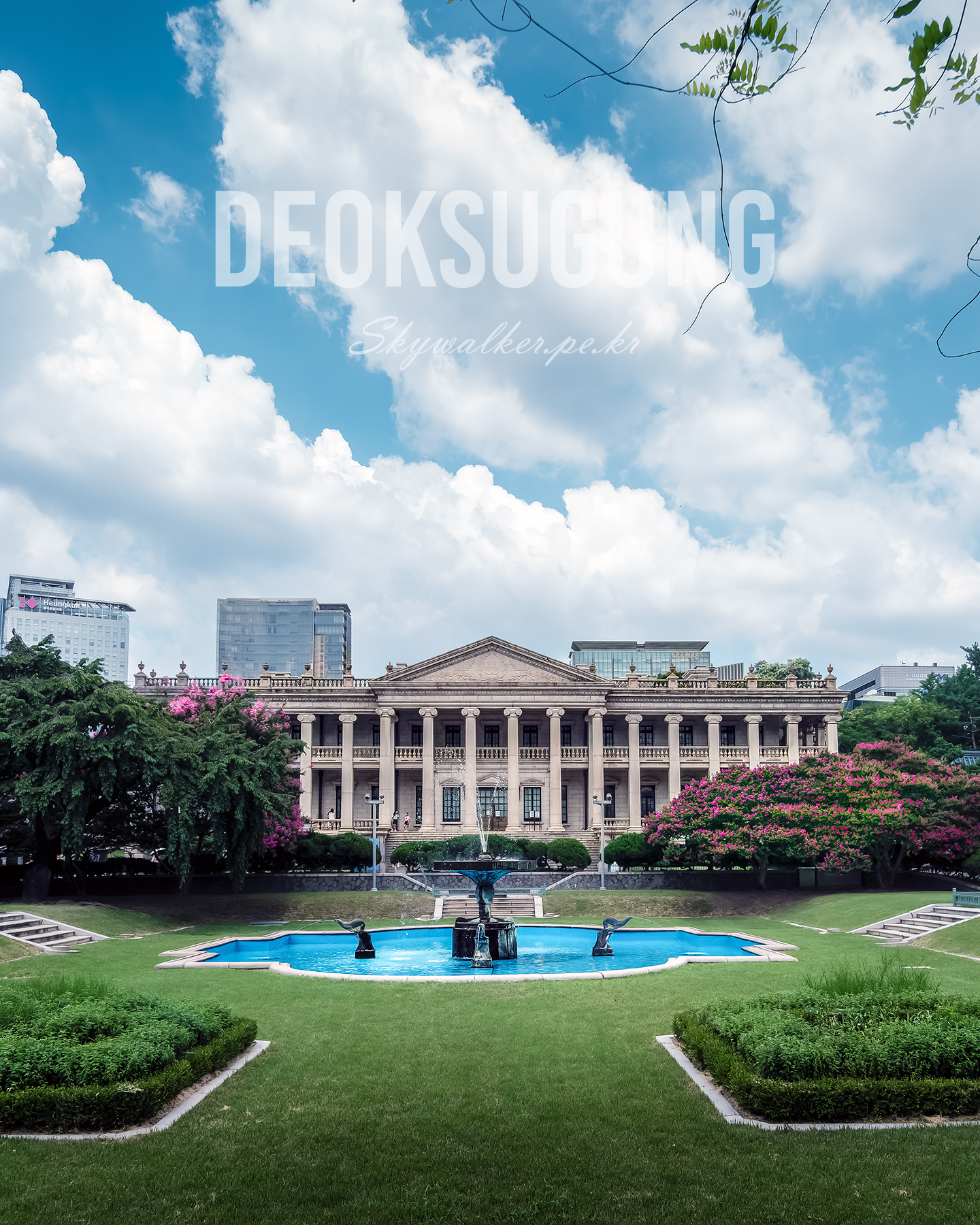 Deoksugung - A classical stone building stands behind a fountain and manicured lawn under bright clouds, framed by flowering trees and city towers. - #Deoksugung #DeoksugungPalace #DeoksuPalace #Seoul #SeoulKorea #SouthKorea #KoreanPalace #KoreanHistory #KoreanHeritage #CulturalHeritage