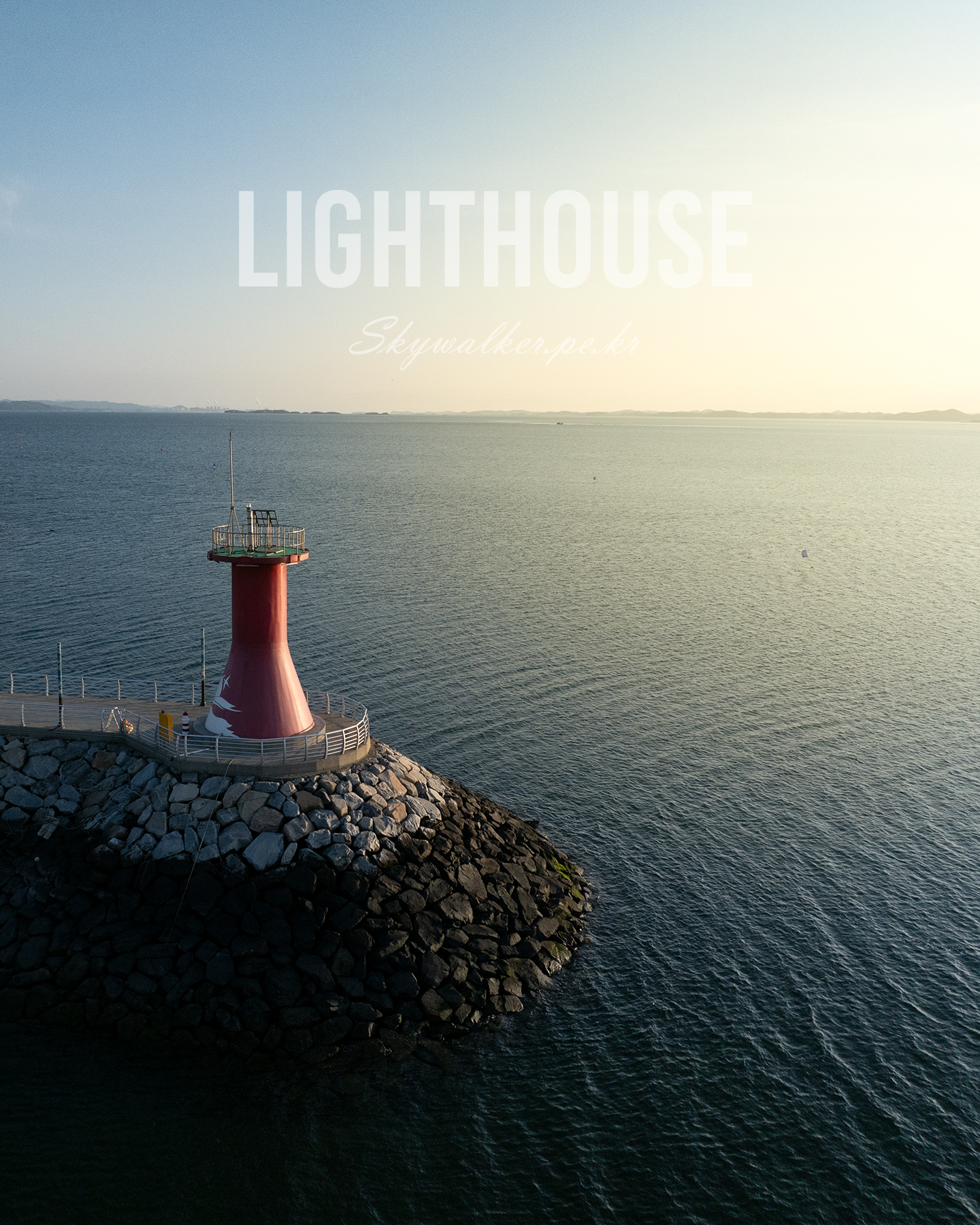 Lighthouse - A red lighthouse stands on a rocky breakwater beside a calm sea, lit by warm low sunlight under a hazy sky. - #Lighthouse #LighthousePhotography #CoastalLandscape #Seascape #OceanView #SeaView #Coastline #CoastalPhotography #Maritime #Seaside