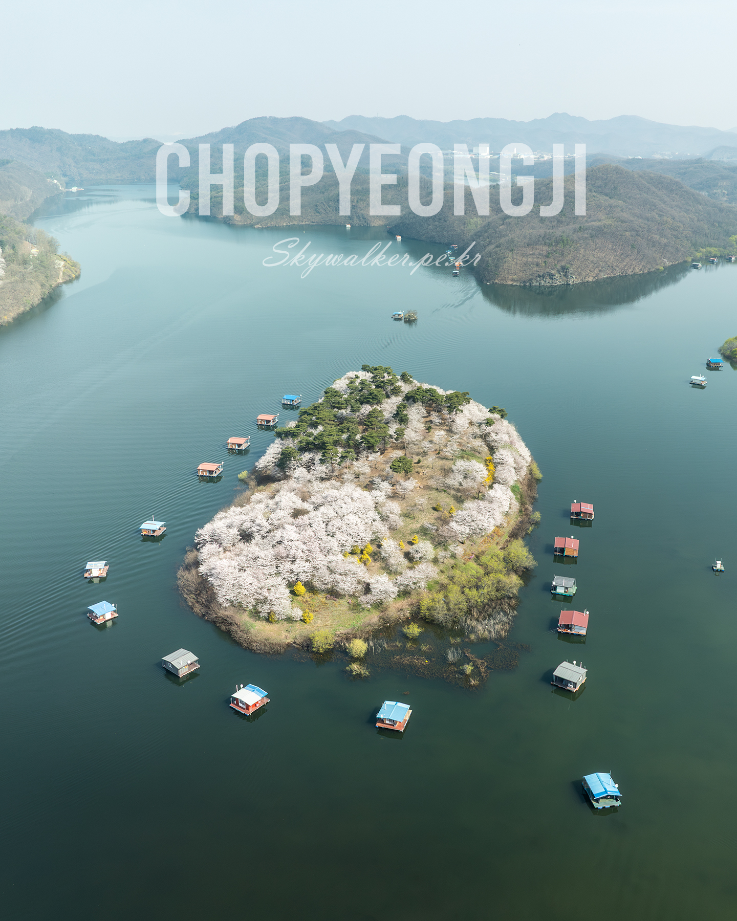 Chopyeongji - An aerial view shows a flowering island surrounded by calm lake water, floating huts, and hazy wooded hills under soft daylight. - #Chopyeongji #ChopyeongReservoir #ChopyeongLake #Jincheon #JincheonKorea #Chungcheongbukdo #KoreaLandscape #LakeLandscape #Lakeside #LakeView