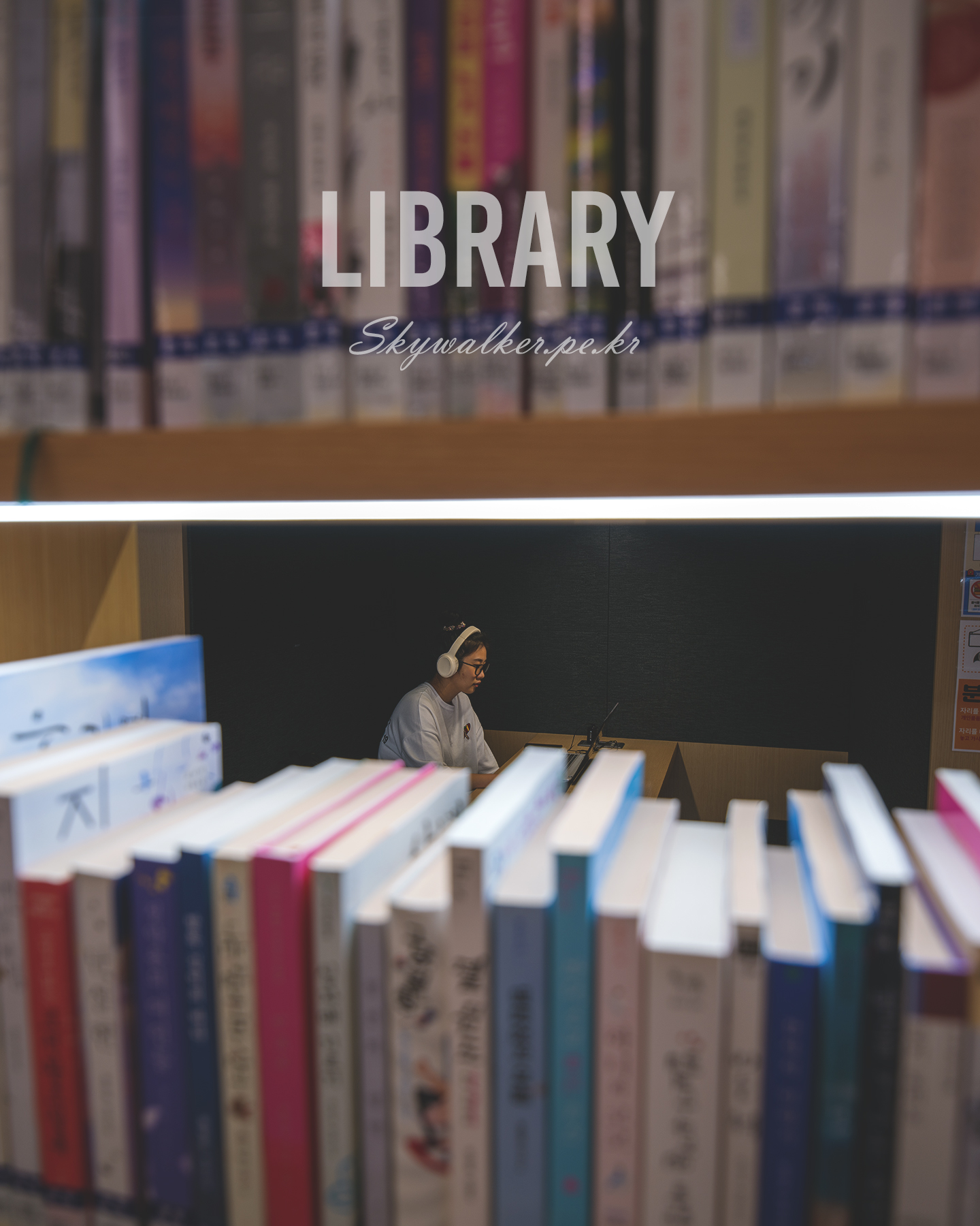 Library - A person wearing headphones works at a laptop in a library, framed by blurred bookshelves and a bright horizontal light. - #Library #LibraryPhotography #PublicLibrary #Books #Bookshelf #BookLovers #ReadingSpace #ReadingTime #StudySpace #QuietMoments
