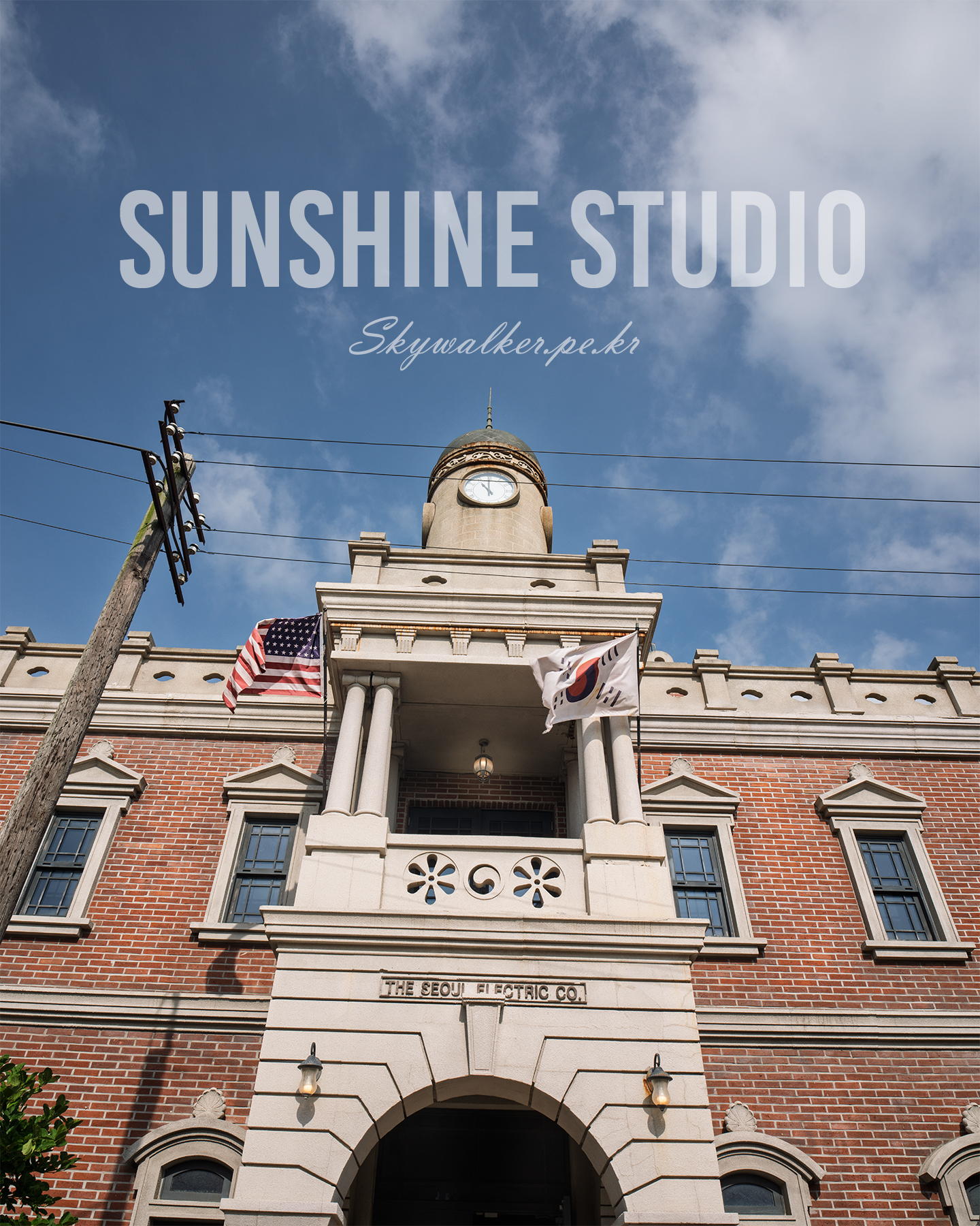 Sunshine Studio - A sunlit red-brick building with a clock tower, flags, and overhead wires stands beneath a blue sky with scattered clouds. - #SunshineStudio #MrSunshine #NonsanSunshineStudio #Nonsan #KDrama #KDramaFilmingLocation #KoreanDrama #DramaSet #FilmingLocation #KoreanHistory