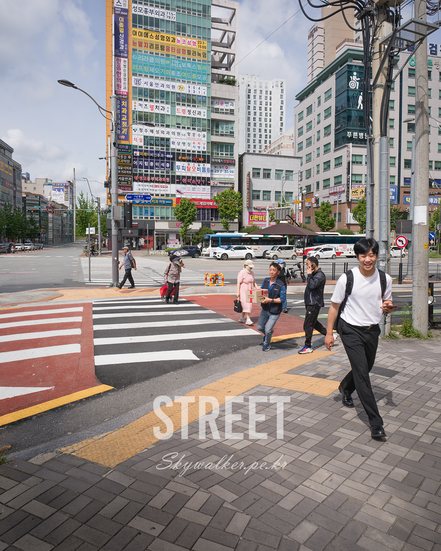 Street - Pedestrians cross a wide urban intersection beneath dense clinic signs and overhead wires in bright midday light. - #Cheongju #CheongjuStreet #CheongjuCity #Crosswalk #PedestrianCrossing #StreetPhotography #UrbanPhotography #StreetScene #CityStreet #CityLife