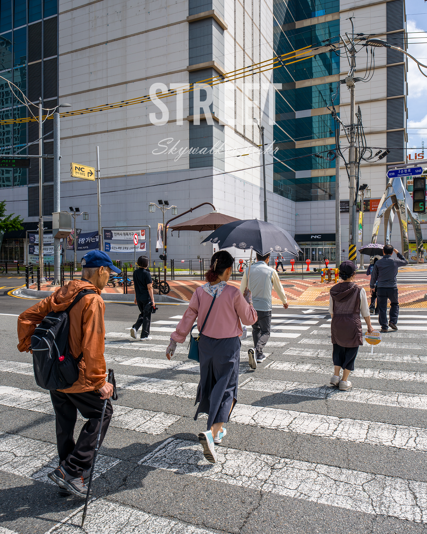 Street - Pedestrians cross a sunlit urban crosswalk beneath overhead wires and modern glass-and-stone buildings. - #Cheongju #CheongjuStreet #CheongjuCity #Crosswalk #PedestrianCrossing #StreetPhotography #UrbanPhotography #StreetScene #CityStreet #CityLife