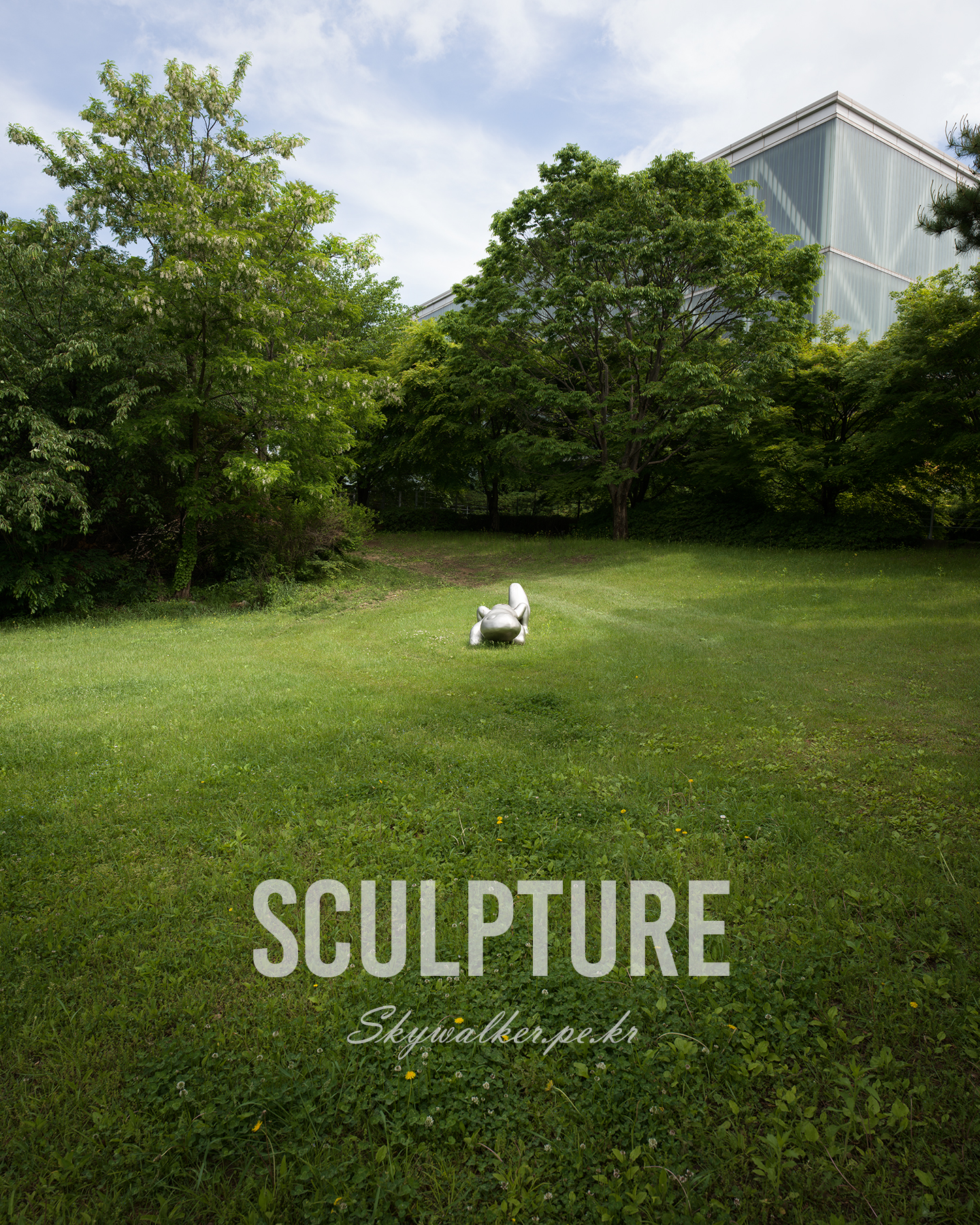 Sculpture - A small silver sculpture rests in a green grassy clearing framed by dense trees and a pale modern building under soft daylight. - #sculpture #publicart #greenlandscape #outdoorart #lushgarden #modernarchitecture #tranquilscene #natureandart #grassyfield #cloudysky
