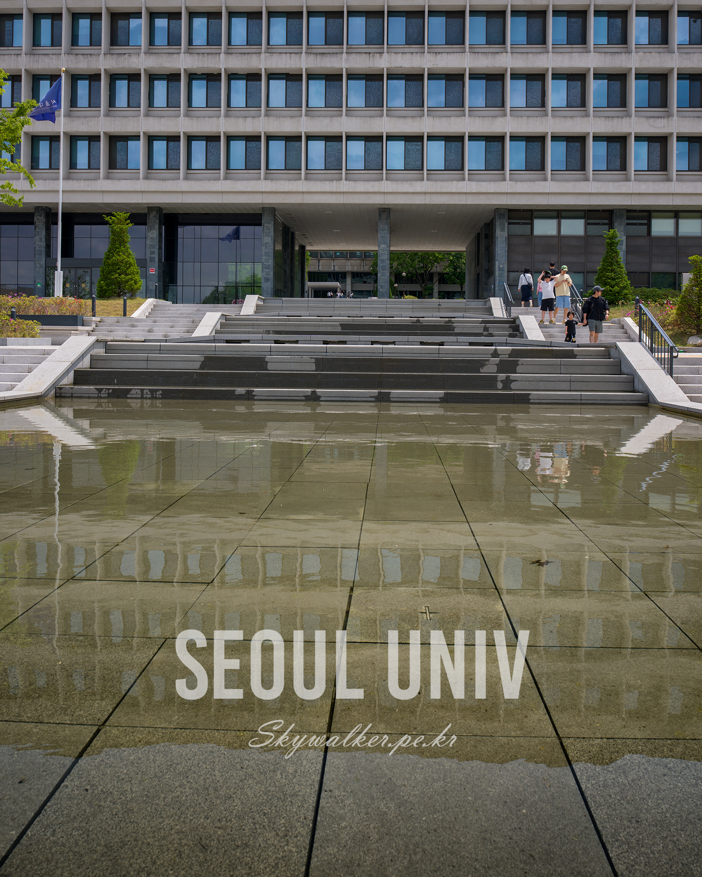 Seoul National University - A modern university building with broad steps is reflected in a shallow water feature under bright daylight. - #CampusArchitecture #ModernArchitecture #ConcreteFacade #ReflectingPool #UrbanCampus #Symmetry #GeometricLines #WetPavement #Stairway #ArchitecturalPhotography