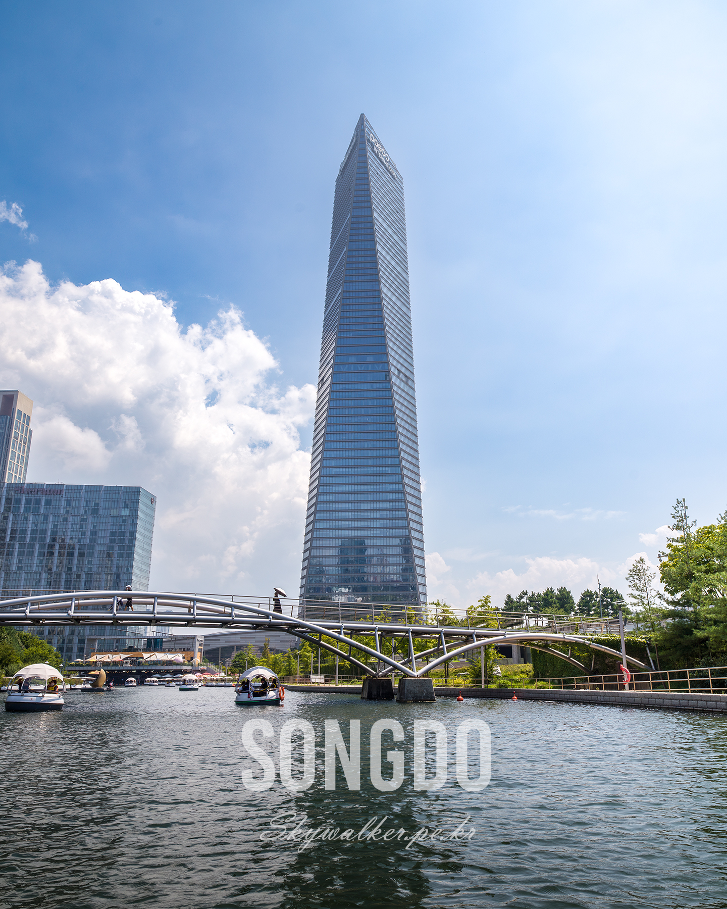Songdo - A tall glass skyscraper rises behind a curved pedestrian bridge and boats on a sunny urban waterway. - #Songdo #Skyscraper #UrbanLandscape #Waterway #Architecture #Cityscape #Bridge #Boats #BlueSky #TravelPhotography