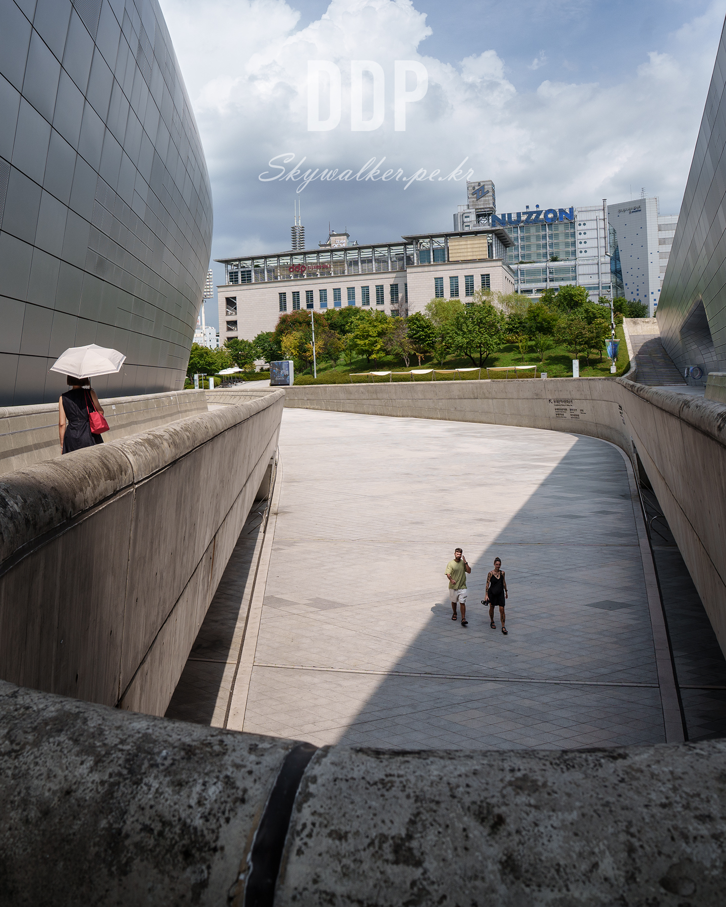 DDP - Pedestrians move through a sweeping concrete walkway between curved metallic buildings under a bright, cloud-filled sky. - #UrbanArchitecture #ModernArchitecture #CurvedLines #ConcreteWalkway #Cityscape #Pedestrians #CloudySky #GeometricComposition #PublicSpace #ArchitecturalPhotography