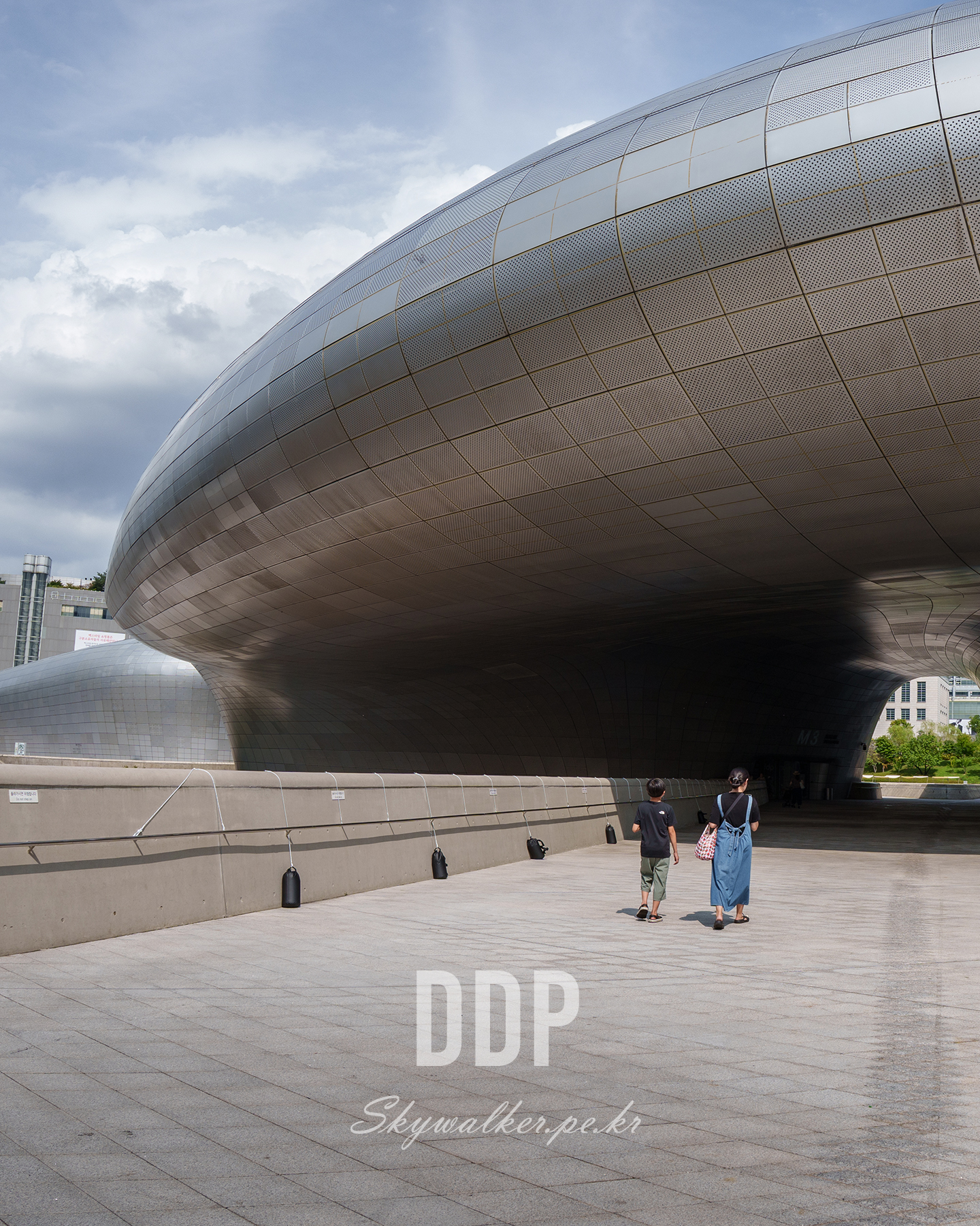 DDP - Two people walk across a broad plaza beneath a sweeping curved metal facade under a bright, partly cloudy sky. - #DDP #architecture #modernarchitecture #urbanphotography #metalfacade #curvedlines #cityscape #cloudysky #publicspace #scale