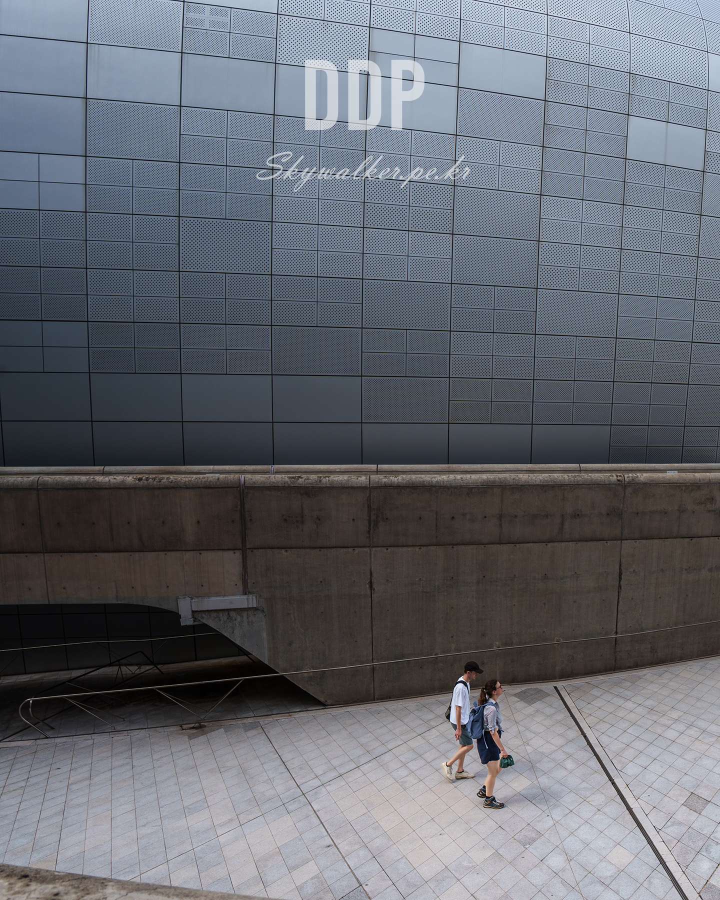 DDP - Two pedestrians walk beneath the patterned metal facade marked DDP, framed by concrete walls and a spacious tiled plaza. - #DDP #Architecture #UrbanDesign #MetalFacade #Concrete #Pedestrians #TiledPlaza #Geometric #Cityscape #Minimalism