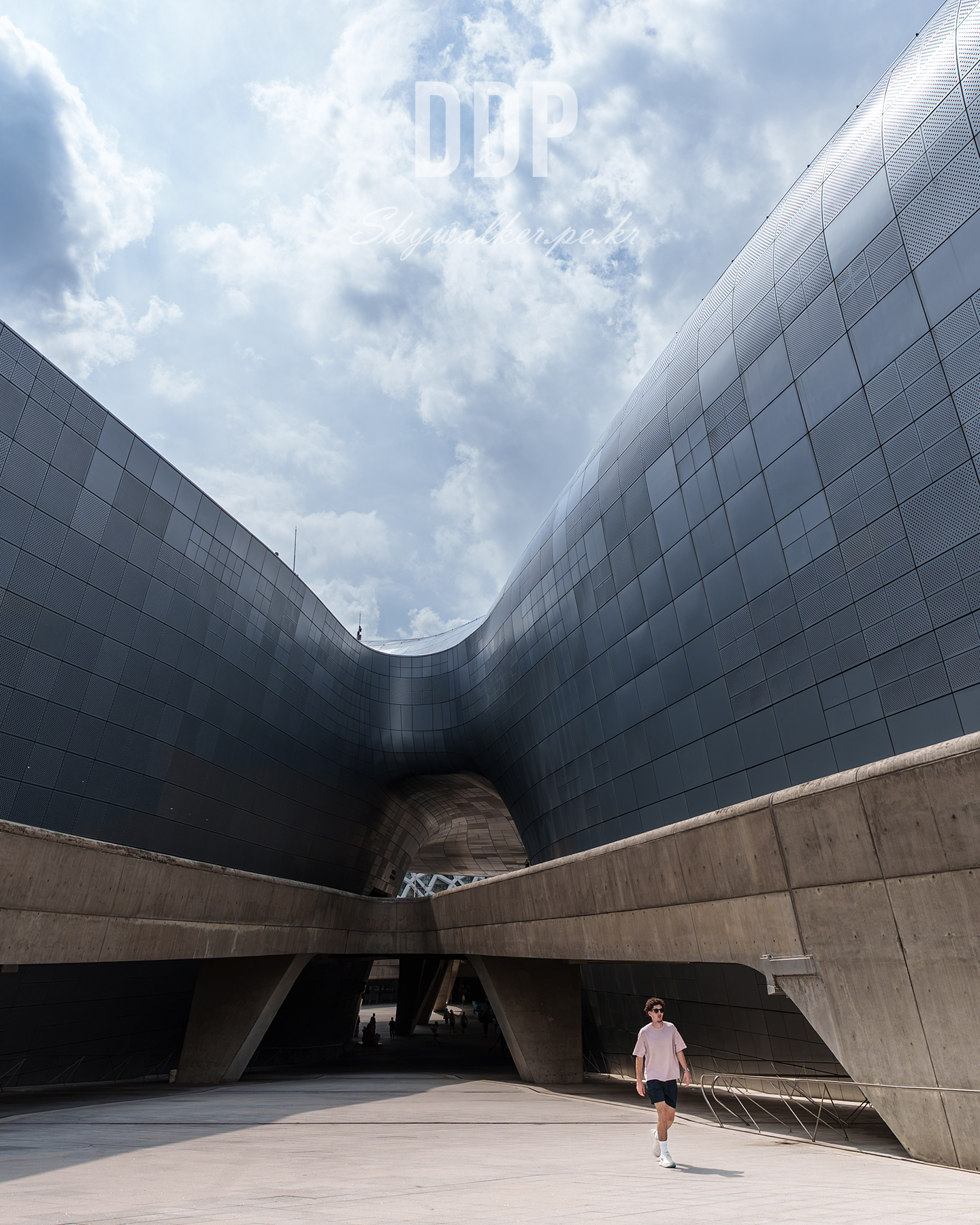 DDP - Curving metallic architecture frames a cloudy sky above a sunlit plaza with a lone pedestrian. - #architecture #modernarchitecture #urbanphotography #metalfacade #curves #cloudysky #perspective #cityscape #minimalism #streetphotography