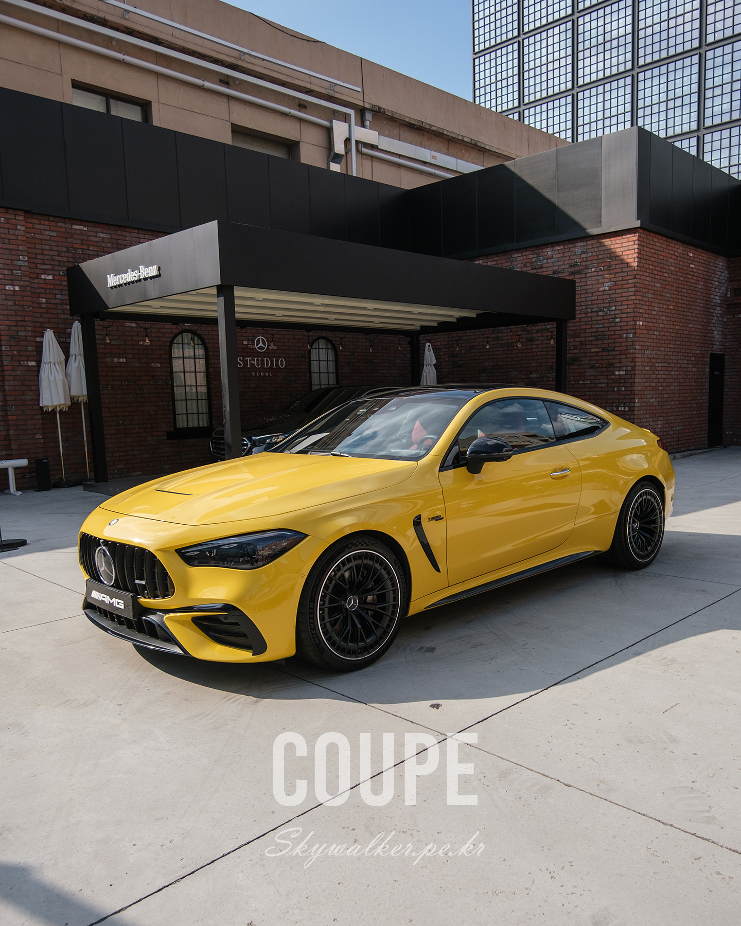 CAR - A yellow Mercedes-AMG coupe is parked in bright sunlight before a brick building with a black modern canopy. - #yellowcar #MercedesAMG #coupe #sportscar #luxurycar #carphotography #urbanarchitecture #brickbuilding #sunlight #automotive