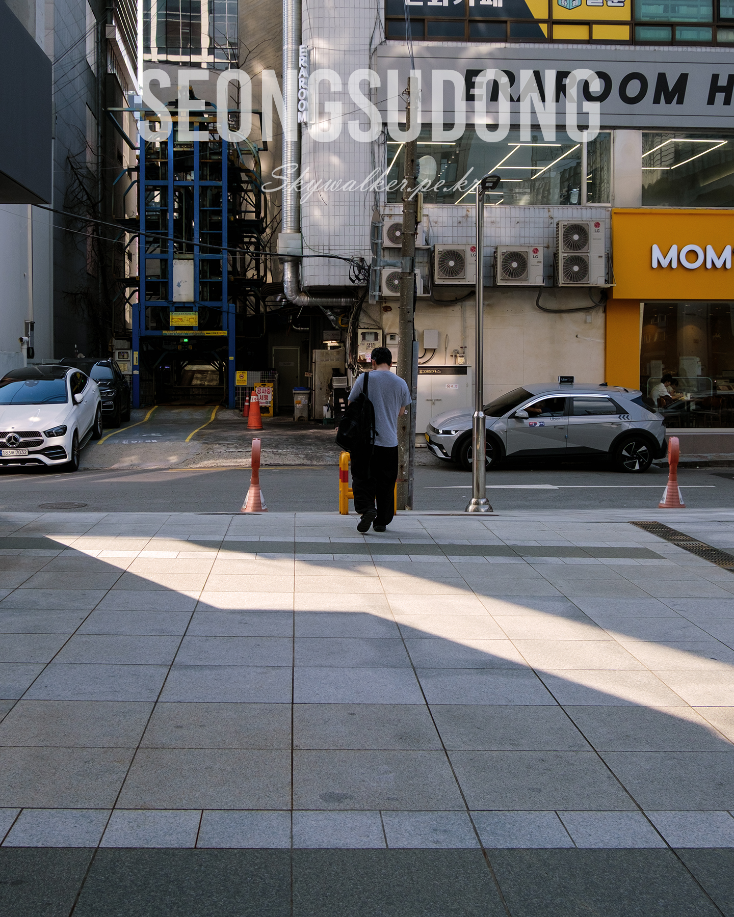 Seongsudong - A lone pedestrian walks along a city sidewalk framed by parked cars, storefronts, and sharp afternoon shadows. - #Seongsudong #StreetPhotography #UrbanScene #CitySidewalk #AfternoonLight #LightAndShadow #Storefronts #ParkedCars #UrbanComposition #EverydayStreet
