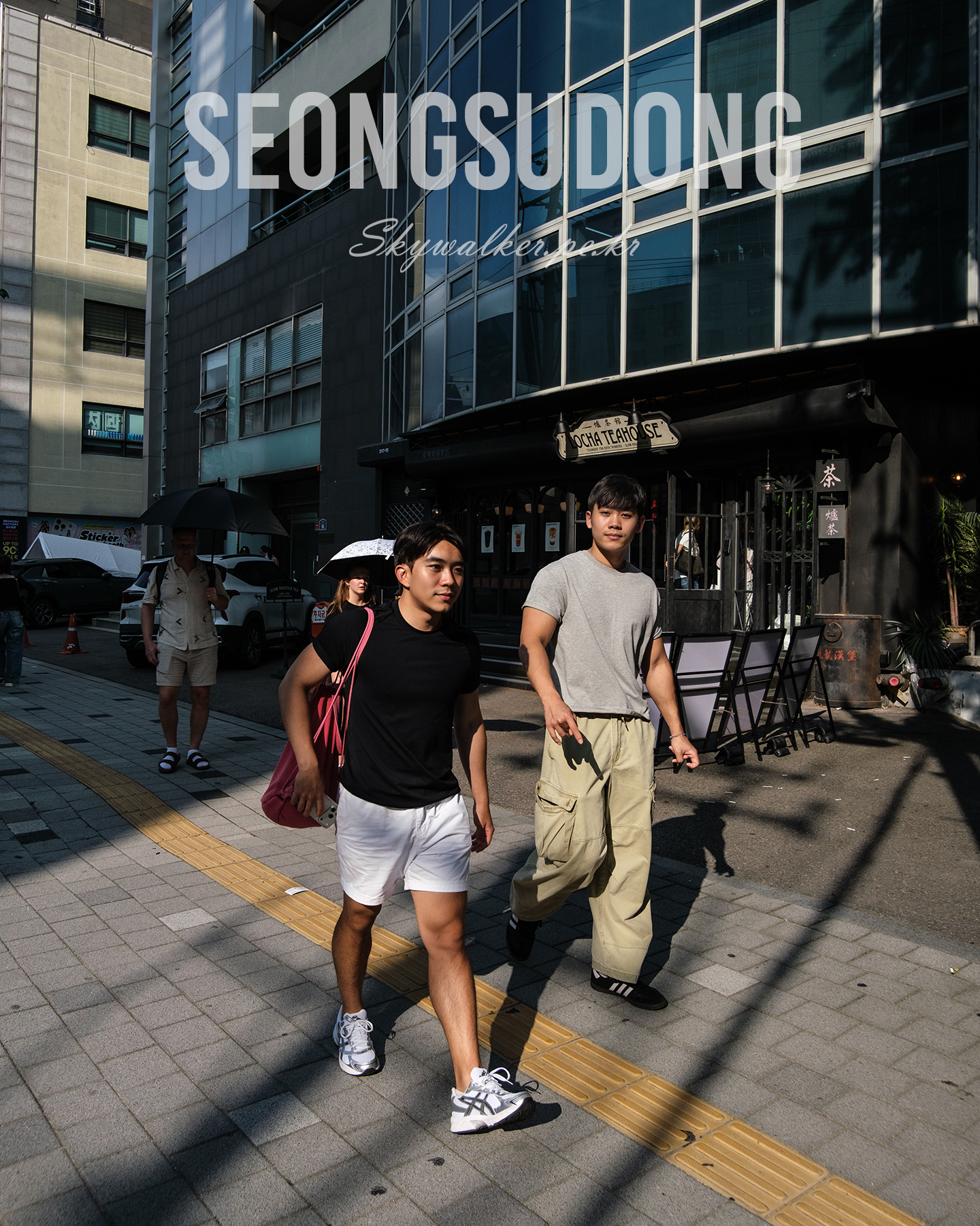 Seongsudong - Pedestrians walk through strong sunlight and deep shadows on an urban sidewalk beside glass-fronted buildings. - #StreetPhotography #UrbanStreet #CityWalk #Pedestrians #Sunlight #Shadows #GlassFacade #Sidewalk #UrbanLife #Seongsudong