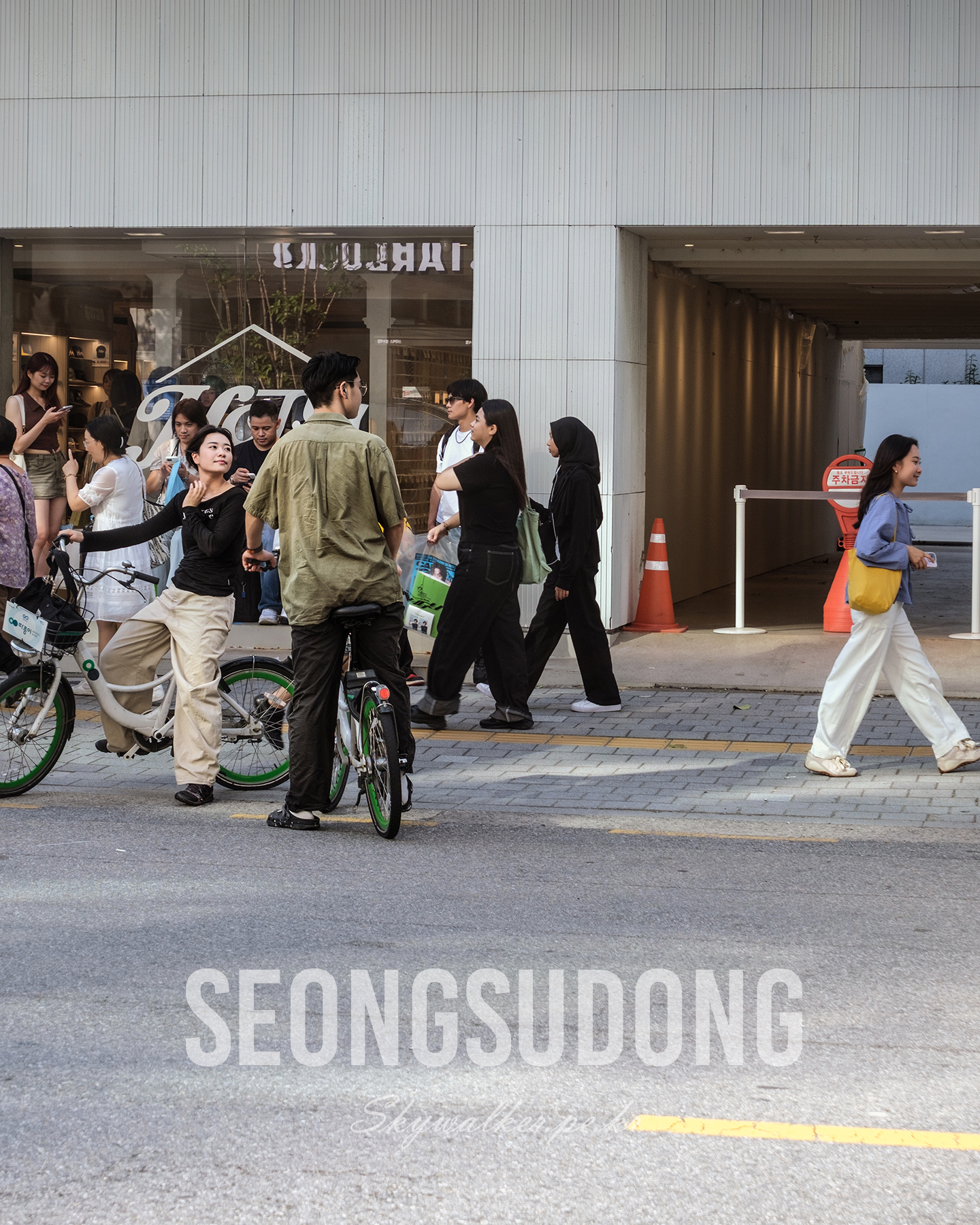 Seongsudong - Pedestrians and people with bicycles gather and pass along a modern storefront and garage entrance in soft daylight. - #StreetPhotography #UrbanLife #Pedestrians #Cyclists #Storefront #CityStreet #Seongsudong #CandidScene #Daylight #Architecture
