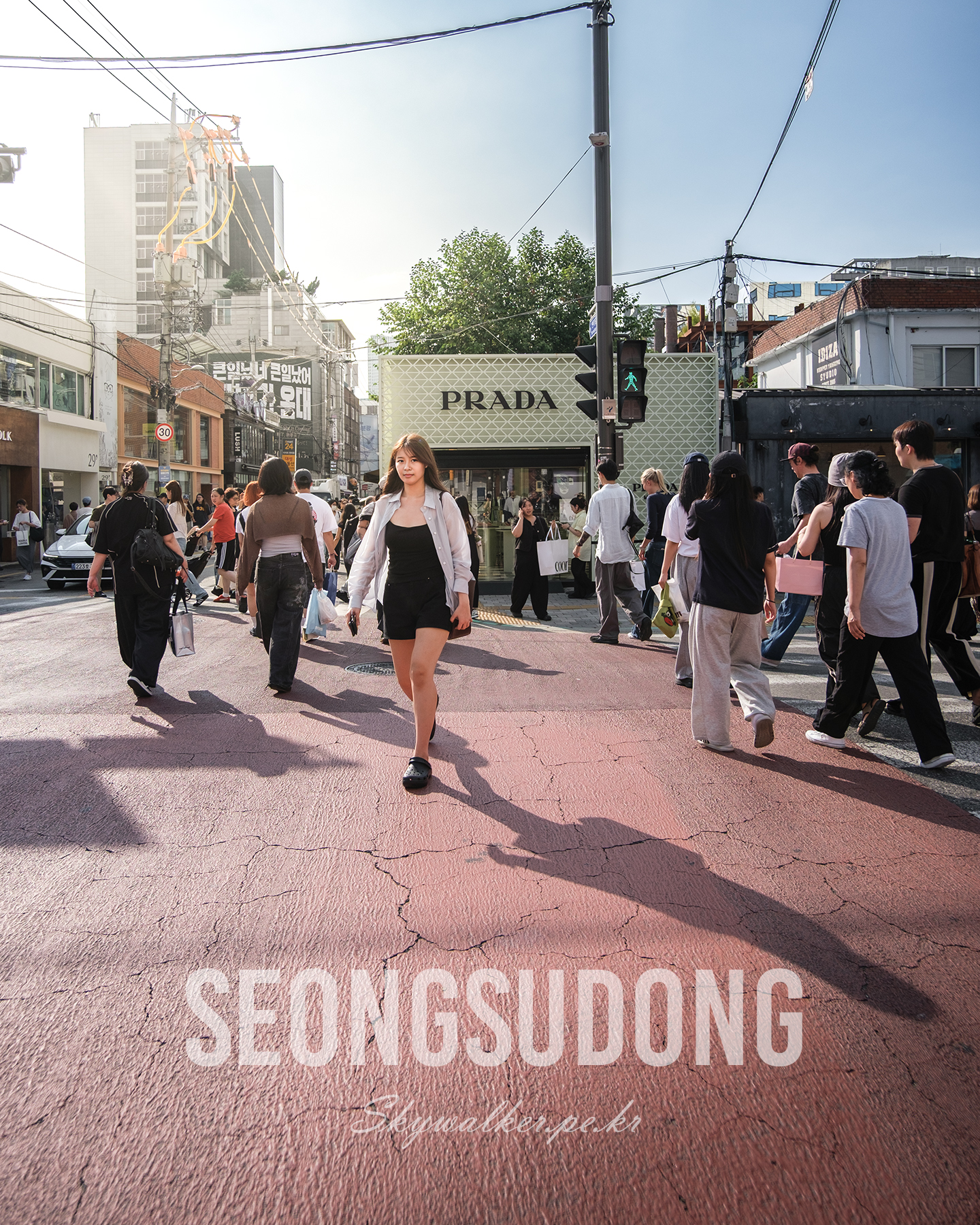 Seongsudong - Pedestrians move through a sunlit urban crosswalk lined with storefronts, overhead wires, and long shadows. - #Seongsudong #StreetPhotography #UrbanScene #CityLife #Pedestrians #Crosswalk #SunlitStreet #LongShadows #Storefronts #OverheadWires