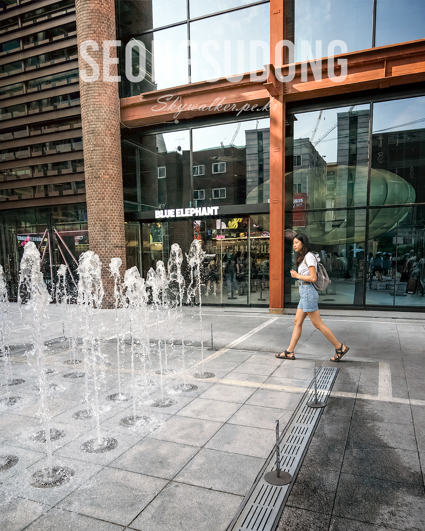 Seongsudong - A woman walks past plaza fountains in front of reflective glass storefronts under warm urban light. - #UrbanPhotography #StreetPhotography #Fountain #CityWalk #GlassFacade #Daylight #ModernArchitecture #Reflection #StreetScene #PublicSpace
