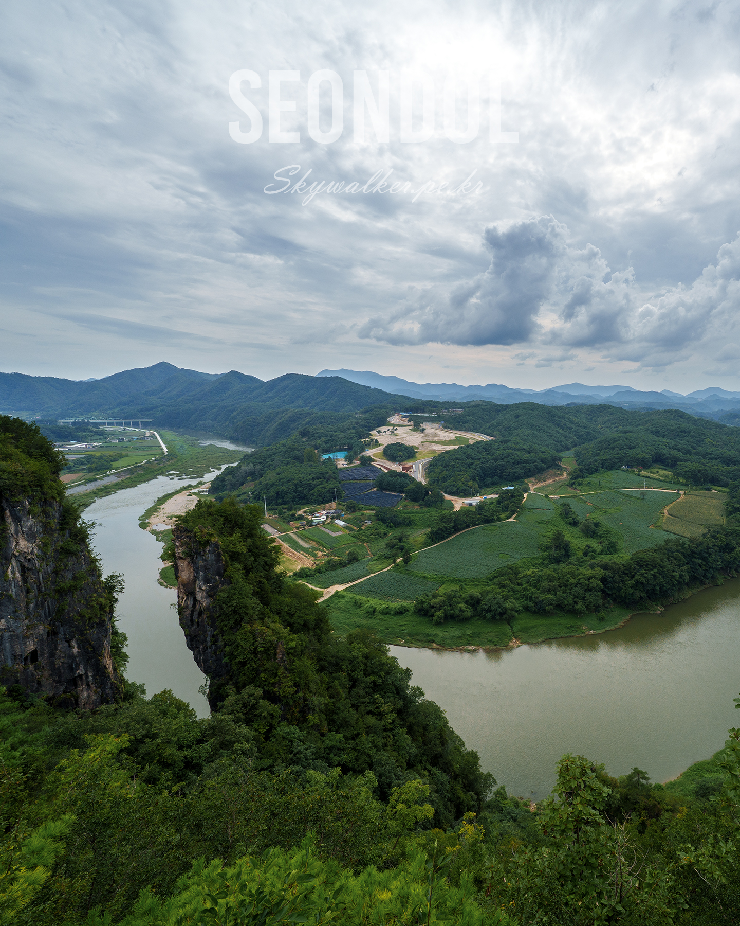 Seondol - A winding river curves through green hills and farmland below rocky cliffs under a dramatic cloudy sky. - #Landscape #River #Mountains #ForestedHills #RockyCliffs #OvercastSky #Panorama #Nature #Valley #ScenicView
