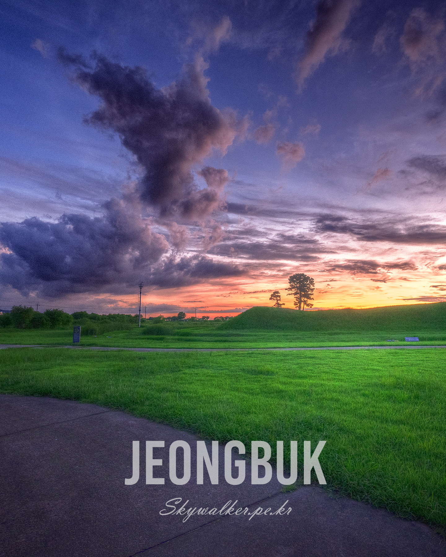 Jeongbuk Fortress - A vivid green field and path sit beneath dramatic purple clouds and a warm sunset, with silhouetted trees on the horizon. - #sunset #cloudscape #landscape #grassland #silhouette #twilight #dramaticSky #greenField #eveningLight #naturePhotography