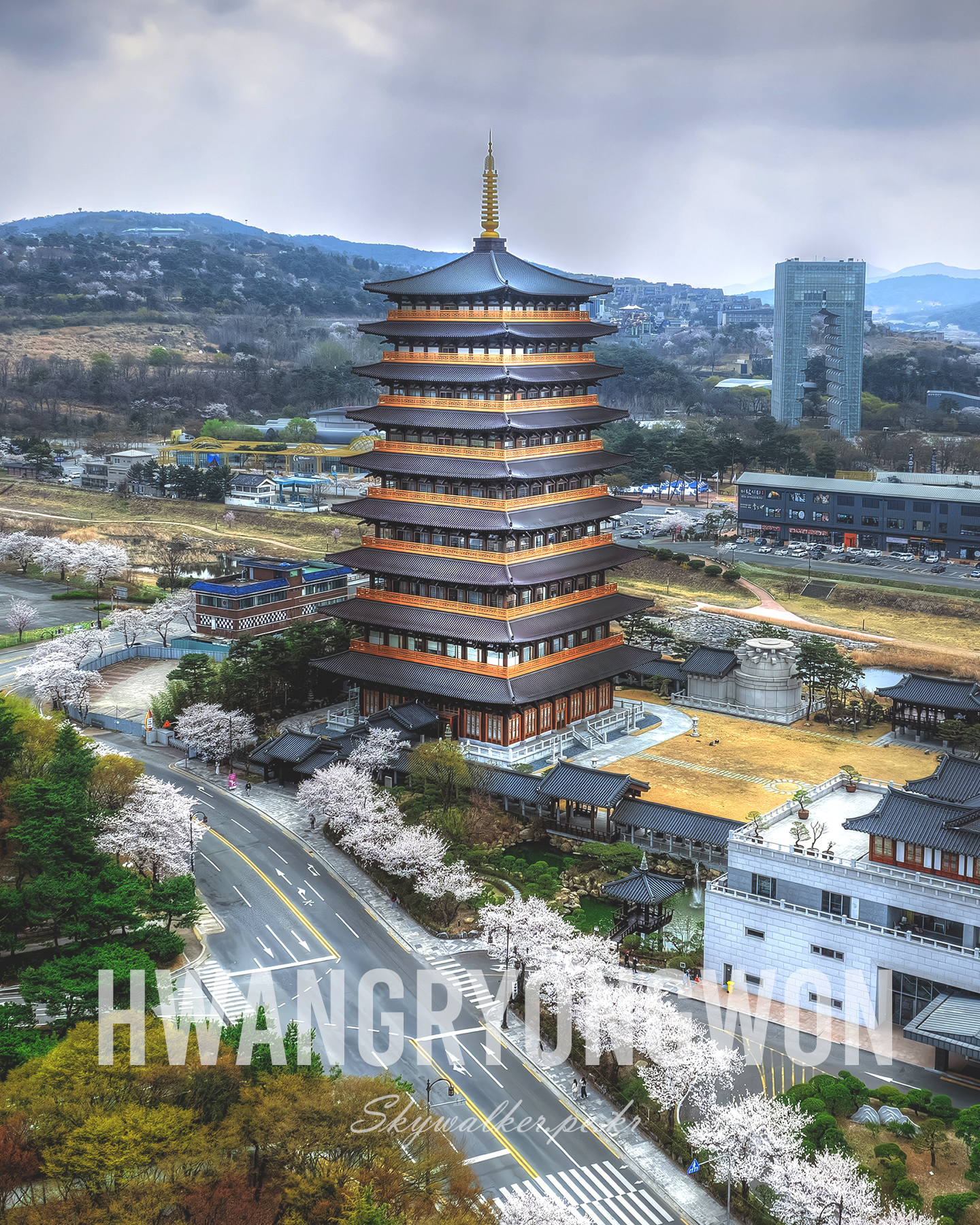 Hwangryongwon - A multi-tiered pagoda rises above cherry blossoms and city streets under soft overcast light. - #Hwangryongwon #Pagoda #TraditionalArchitecture #CherryBlossoms #KoreanArchitecture #CloudySky #AerialView #SpringScenery #UrbanLandscape #LandscapePhotography
